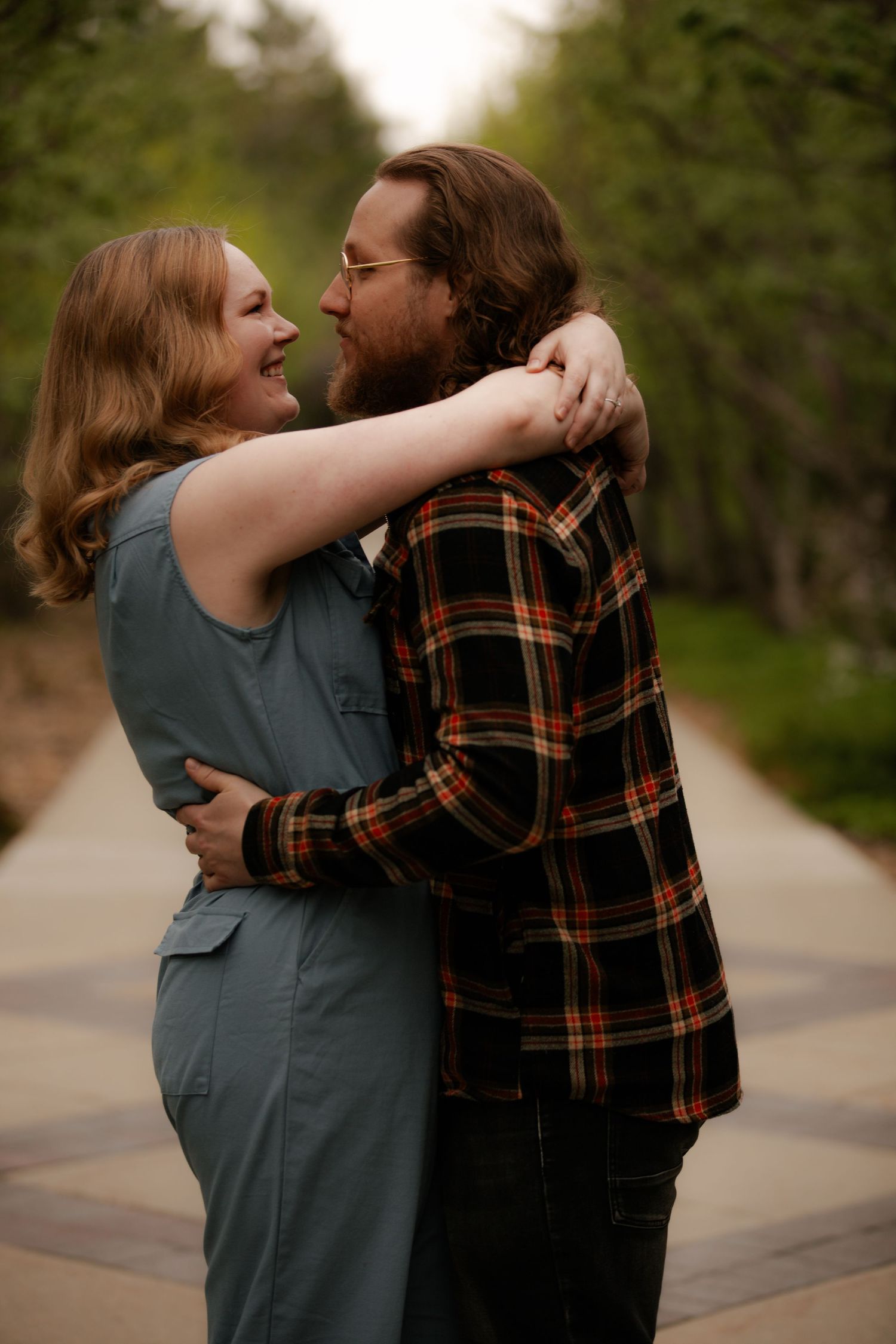 A couple embraces and smiles on a tree-lined path, the woman in a gray jumpsuit hugging a bearded man in plaid.