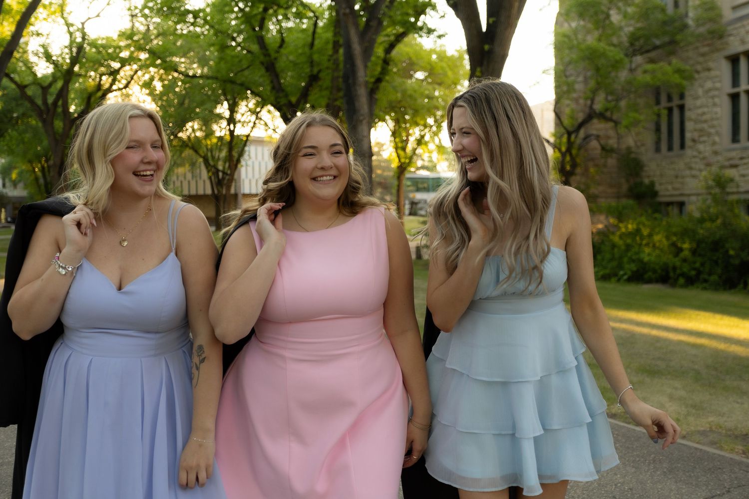 Three young women in pastel formal dresses laughing and walking together outdoors near a stone building.