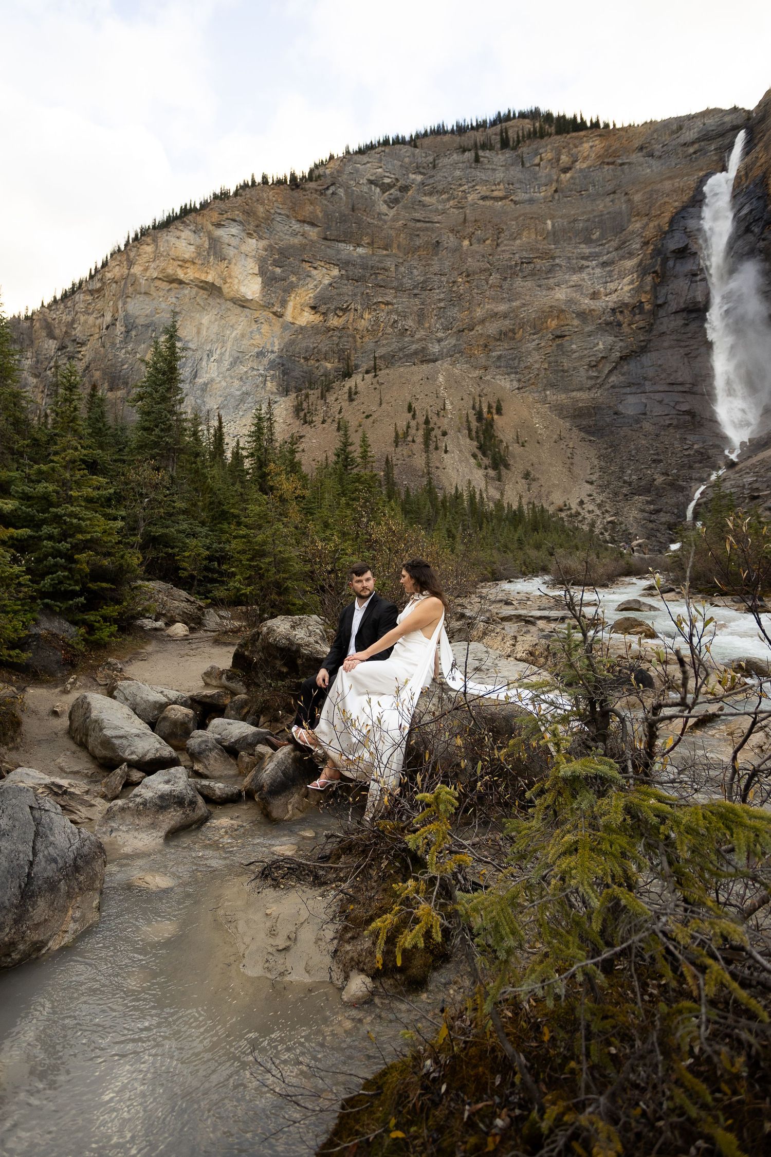 Bride and groom sit on rocks near a mountain waterfall surrounded by evergreen trees in a scenic wilderness setting.