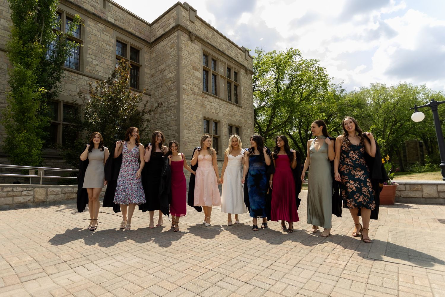 A group of women in elegant dresses walk together outside a stone building on a sunny day.