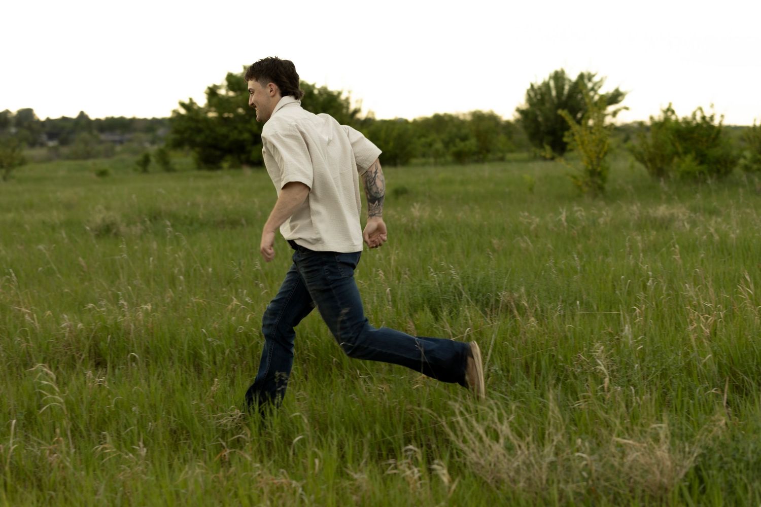 A young man in jeans and white shirt running through a lush green meadow with trees in the background.