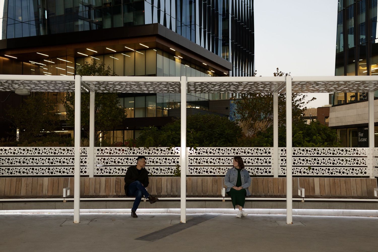 Two people sit at a modern urban bus stop with decorative white panels and glass office buildings in the background.