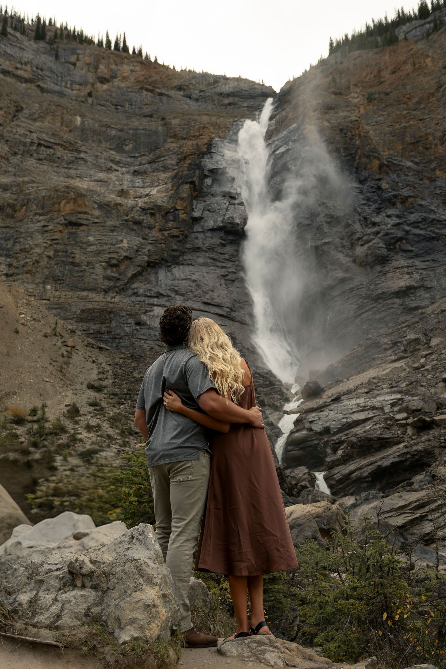 A couple embraces while admiring a majestic waterfall cascading down rocky cliffs in a mountainous landscape Takakkaw Falls in BC, Canada.