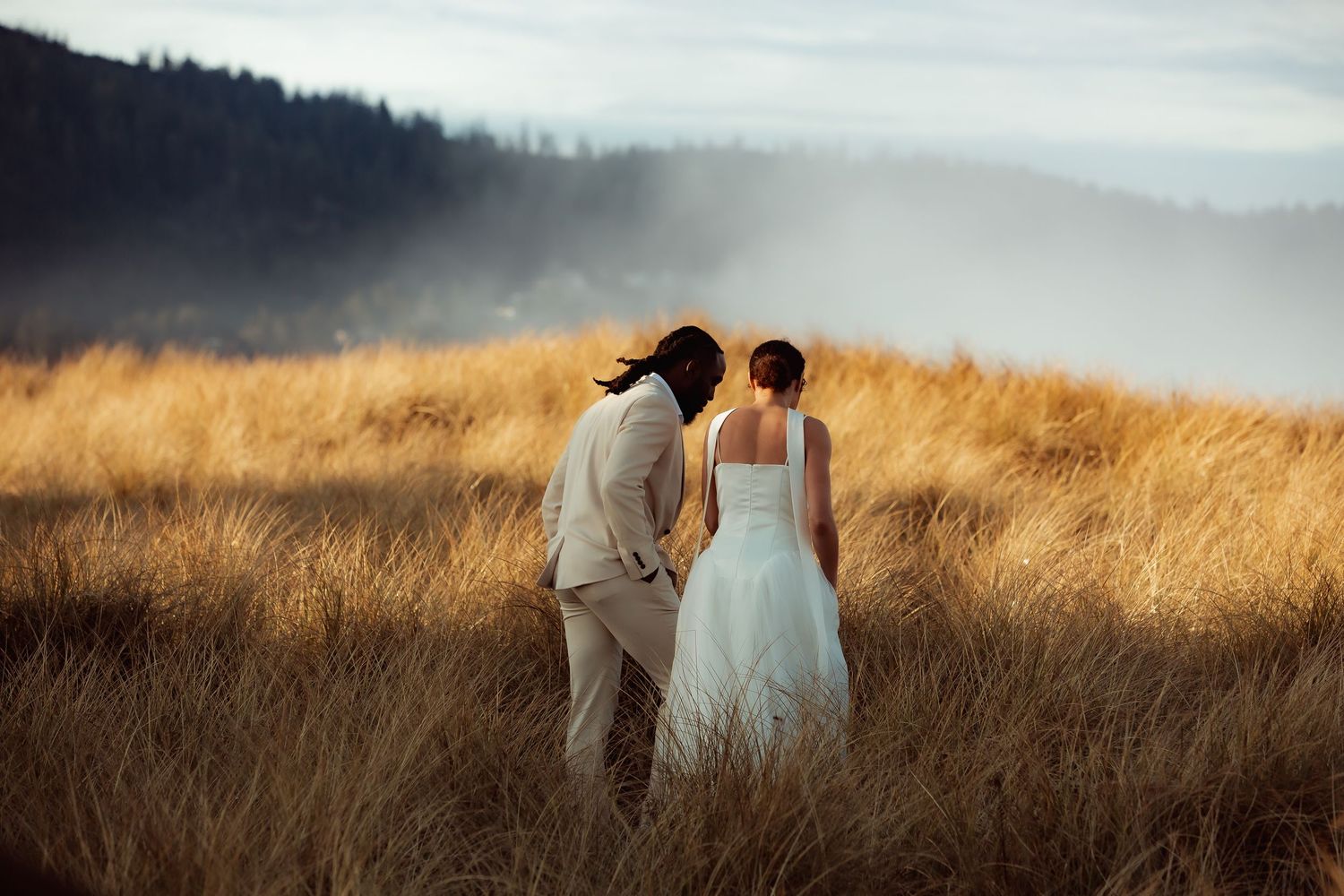A couple in light colored attire walks through a golden wheat field at sunset creating a romantic silhouette.