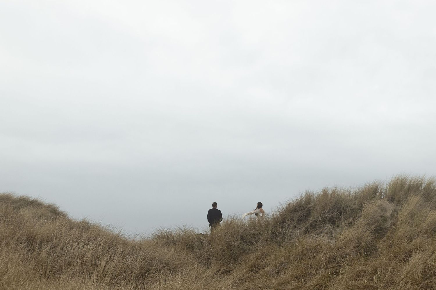 Two people stand among tall grass on coastal sand dunes under an overcast sky.