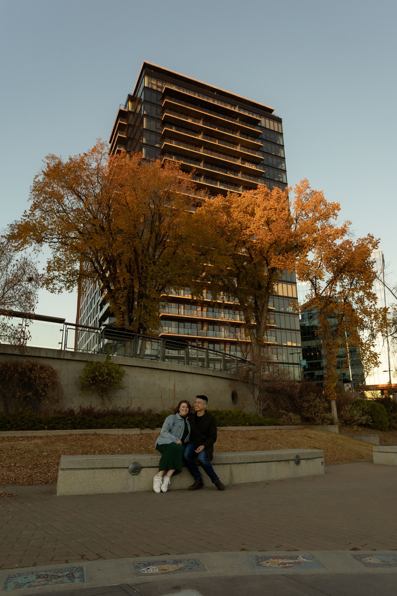 Couple sitting on a bench in an urban plaza with autumn trees and a modern glass tower at golden hour.