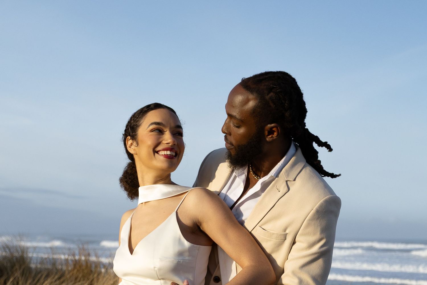 A smiling couple in elegant white attire embraces on a beach with ocean waves and blue sky behind them.