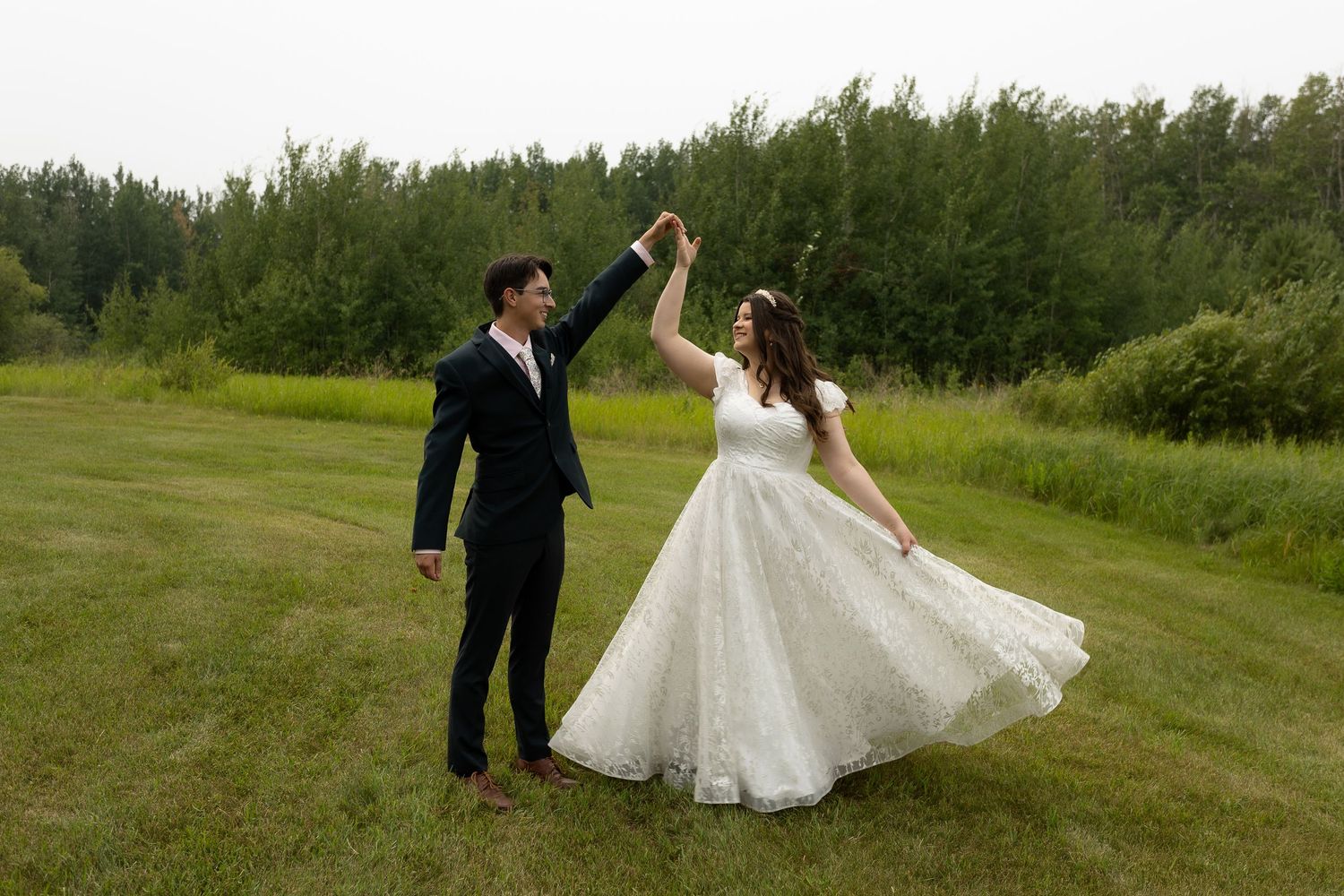 A couple dances joyfully on a grassy field with their wedding attire flowing in the breeze.