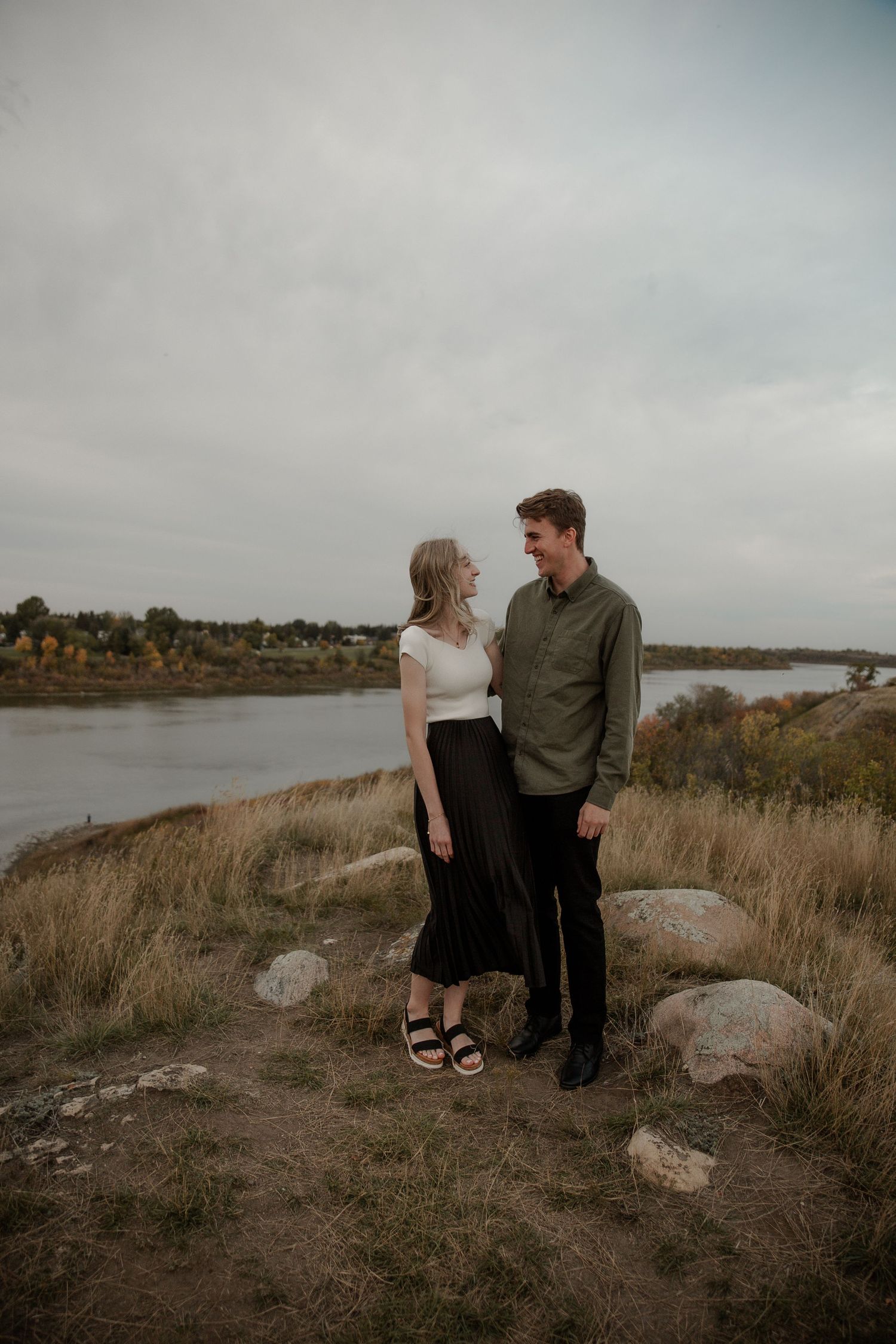 Couple standing together on a rocky hillside overlooking a river, woman in white top and black skirt, man in green shirt.