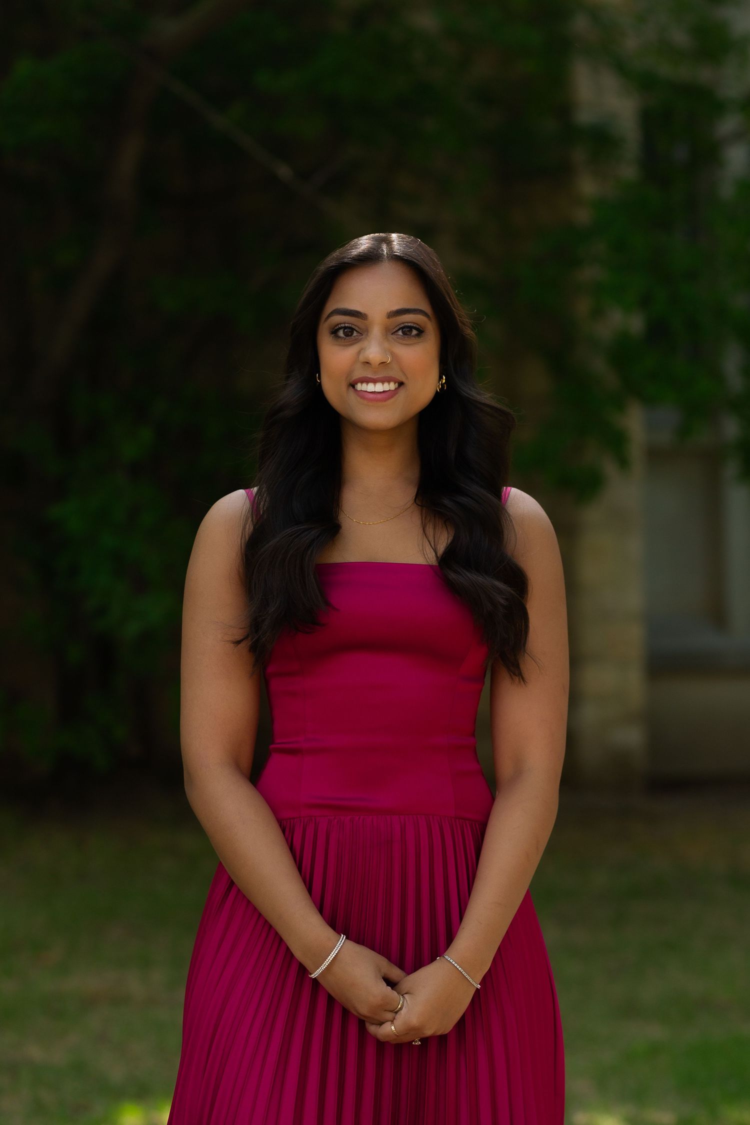 Young woman in magenta pleated dress smiling outdoors with lush green trees in background.