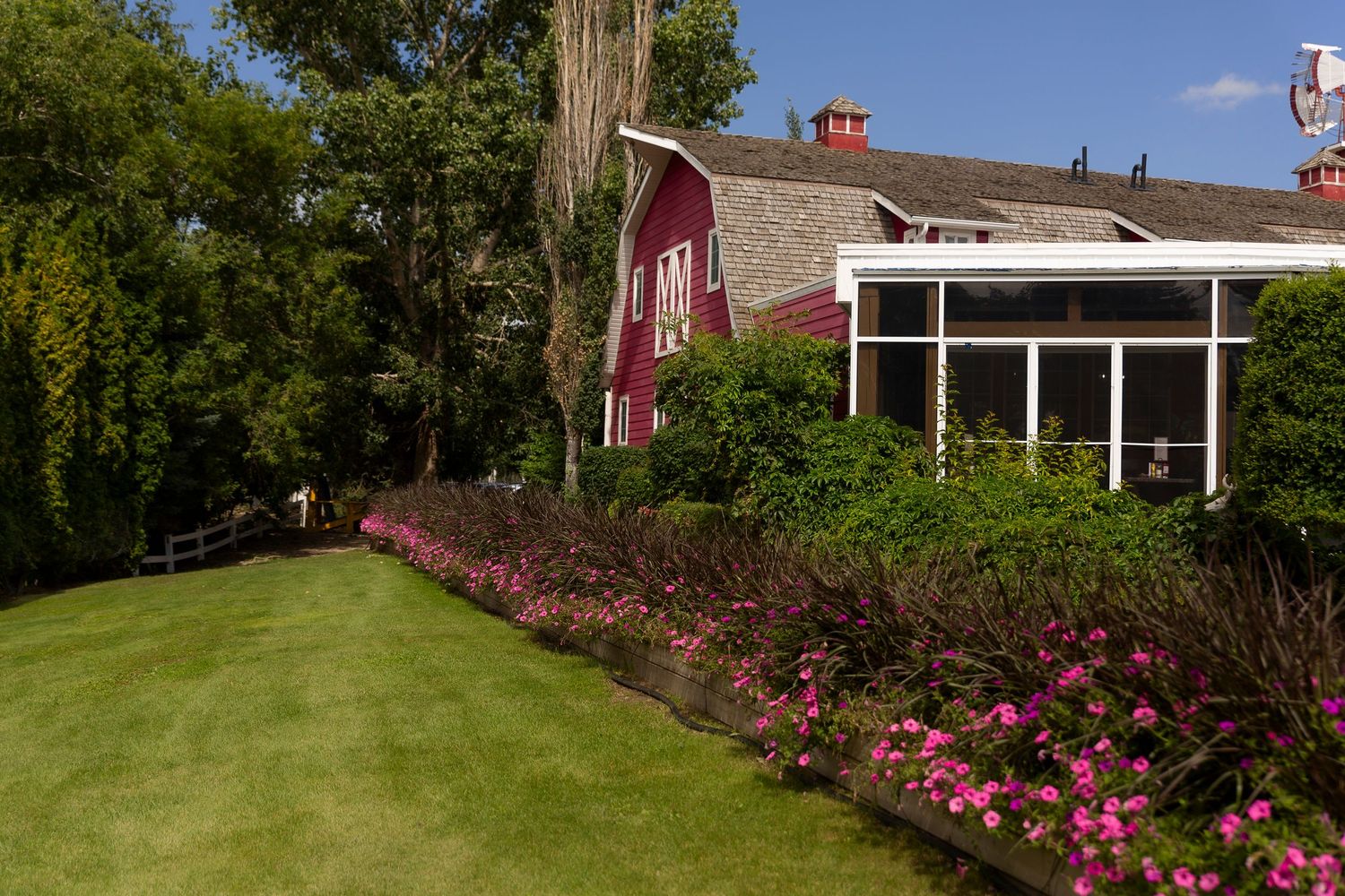 Red barn-style home with screened porch surrounded by lush green lawn and pink flower garden border under blue sky.