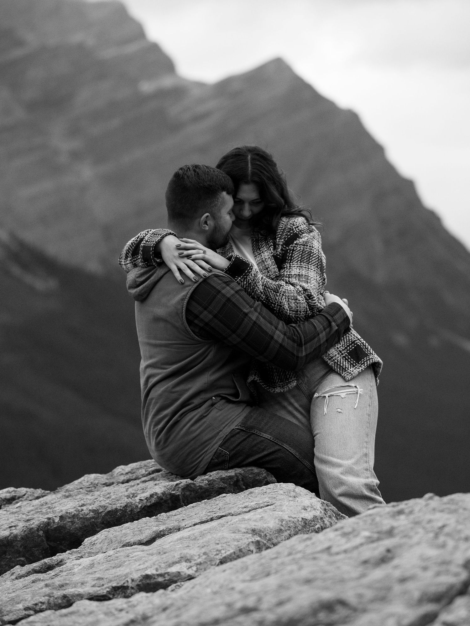 Black and white photo of couple sitting on rocky mountain ledge, sharing an intimate embrace with peaks in background.