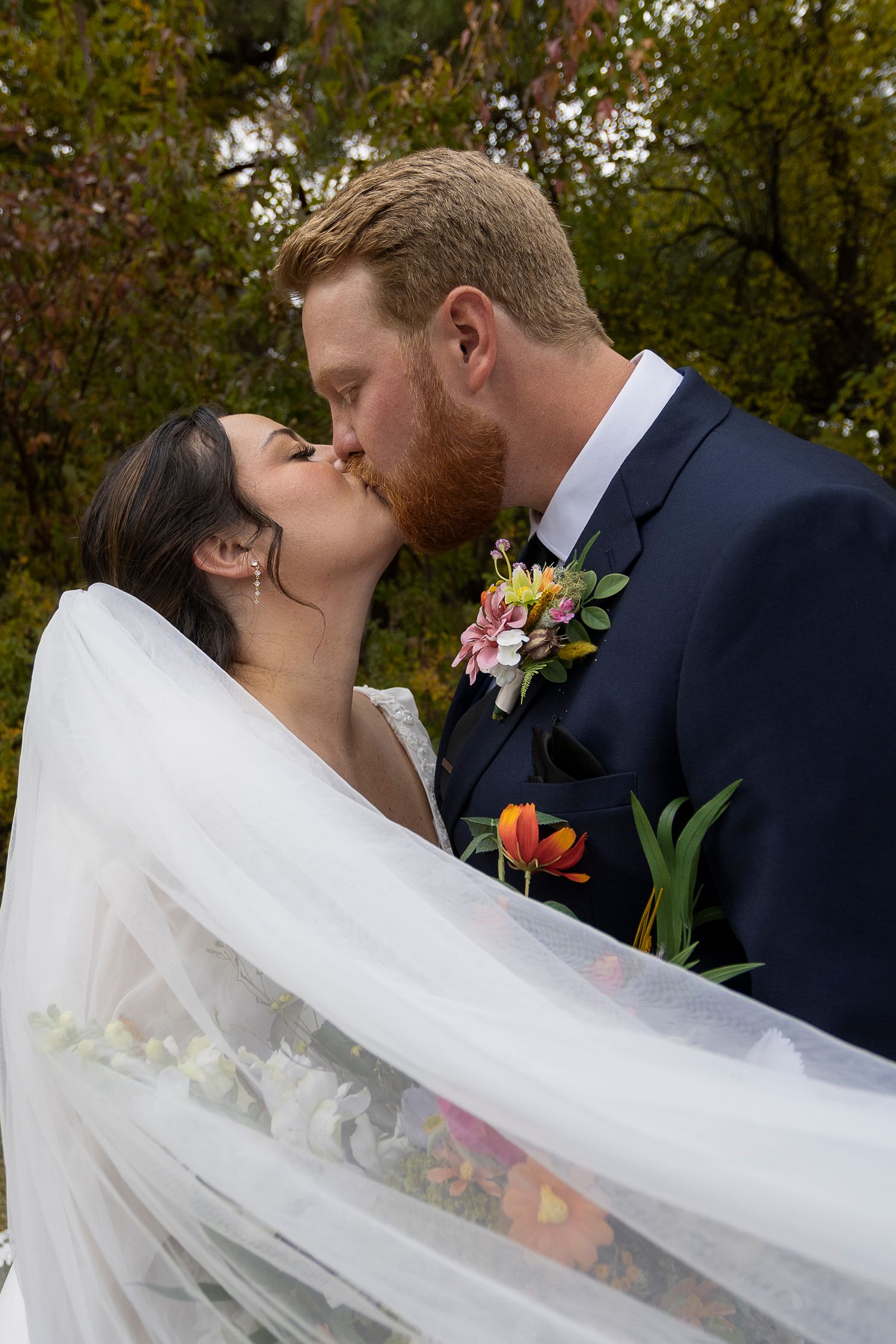 Bride and groom sharing a romantic kiss outdoors, with flowing white veil and colorful floral bouquet and boutonniere.