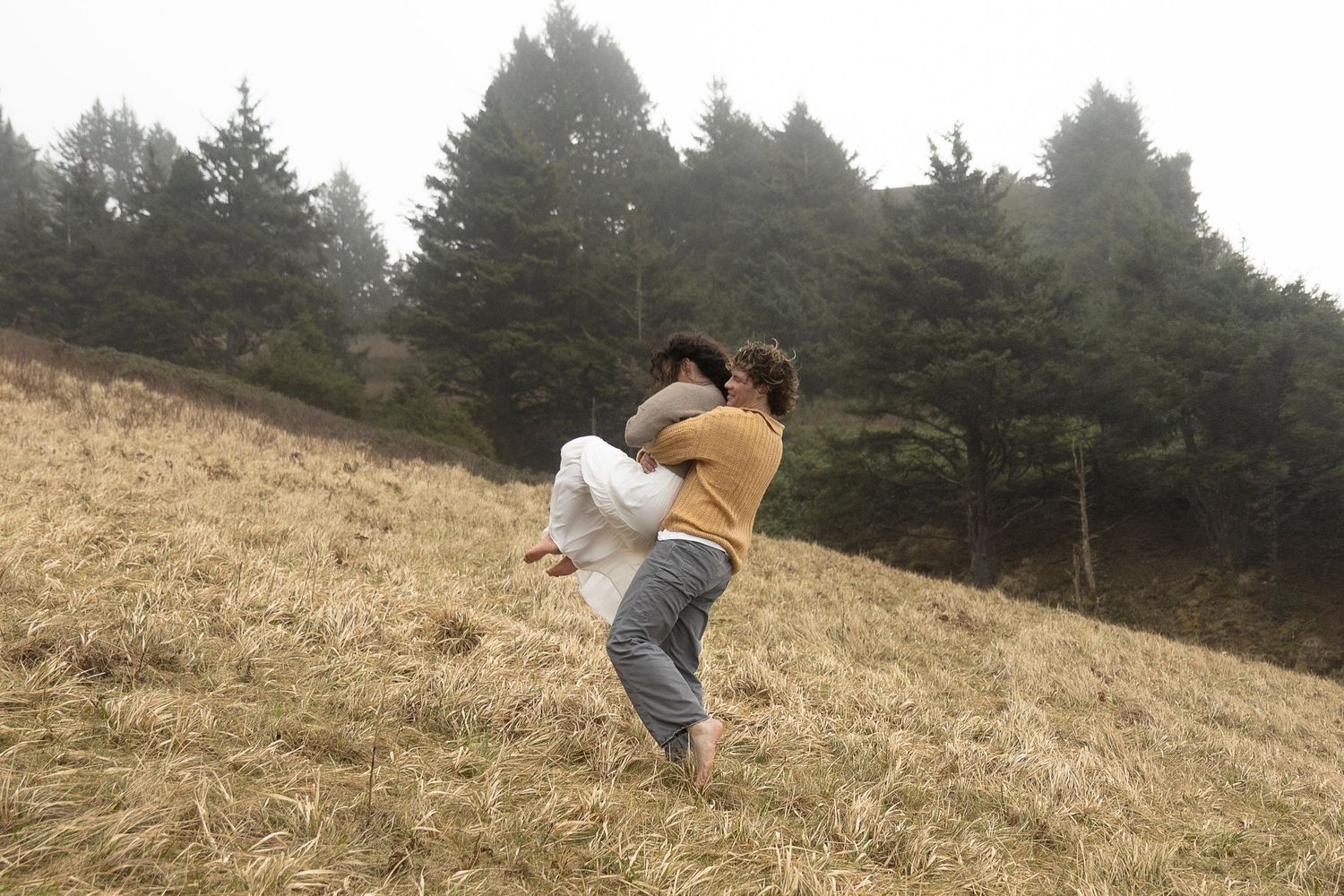 Playful couple embracing on a grassy hillside with evergreen trees in the background.