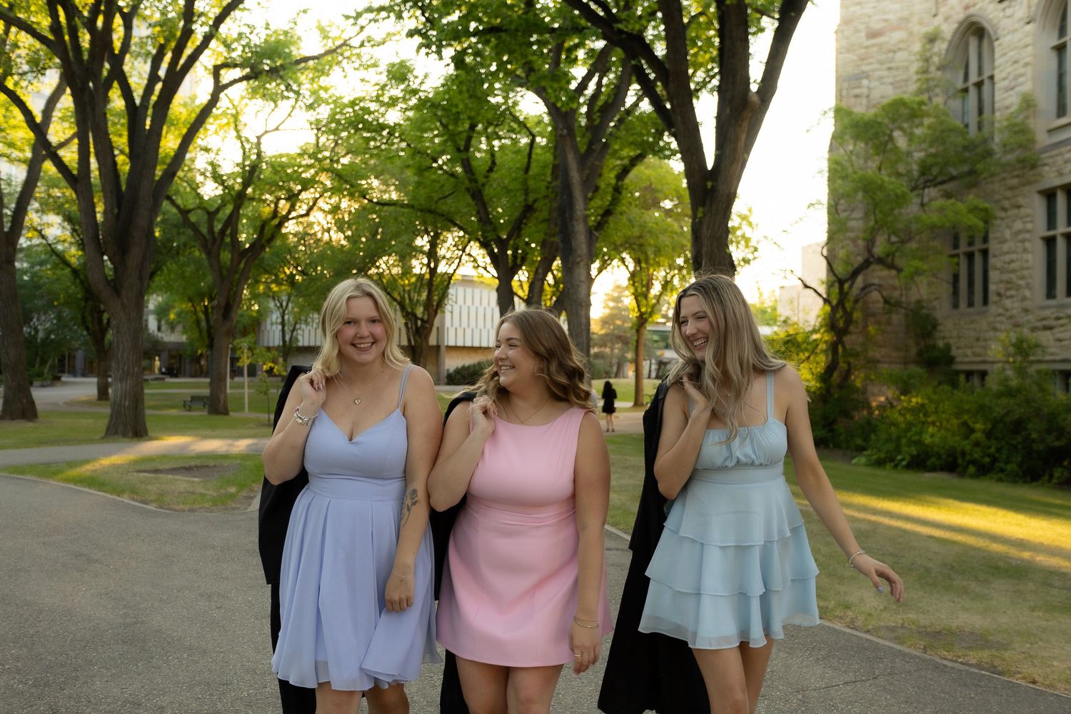 Three young women in pastel dresses walking together outdoors on a tree-lined campus path.
