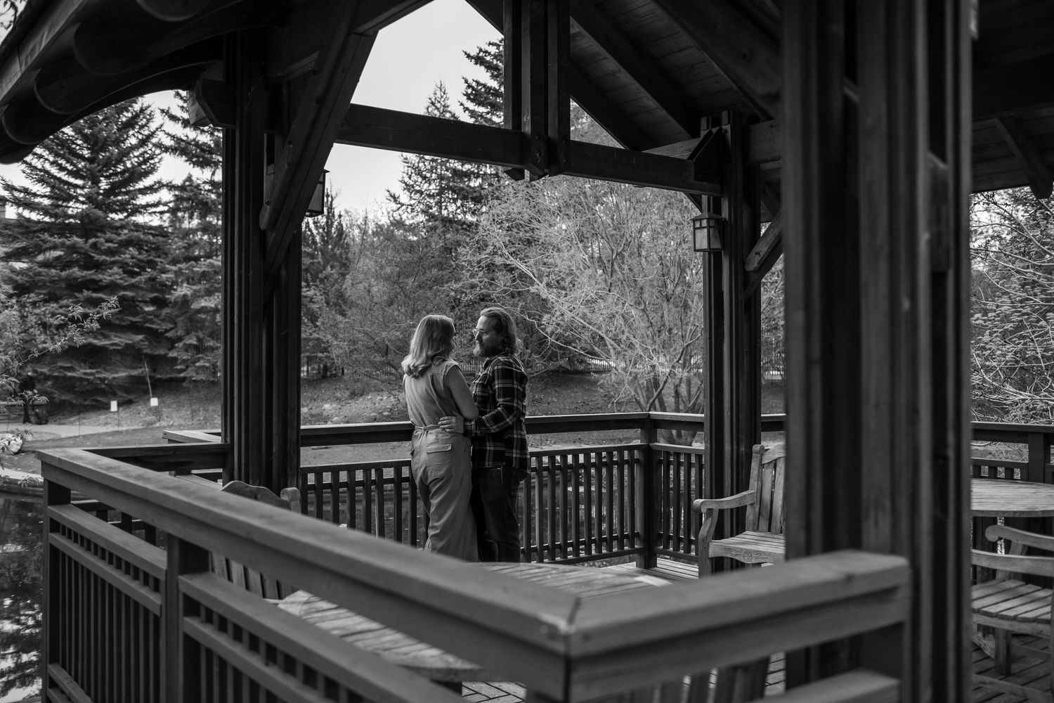 Two people stand on a rustic wooden gazebo overlooking a serene tree-lined landscape in this black and white photo.