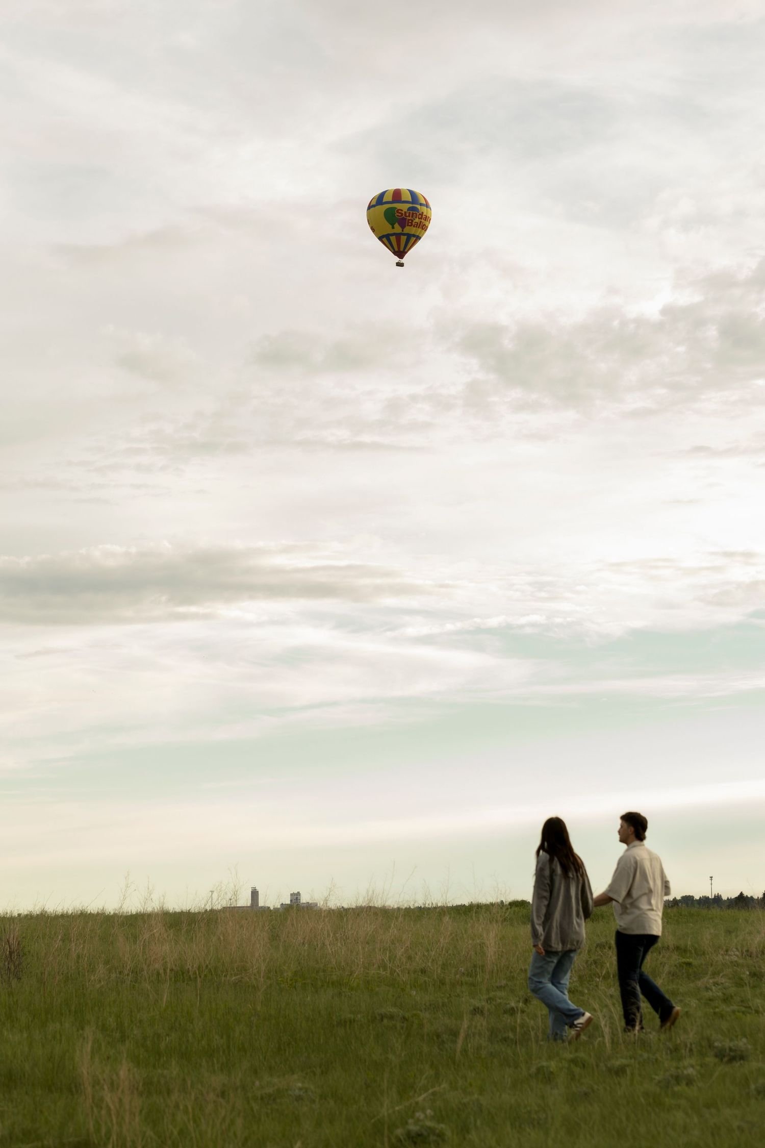 Couple walking hand in hand through green field watching colorful hot air balloon float in cloudy sky.