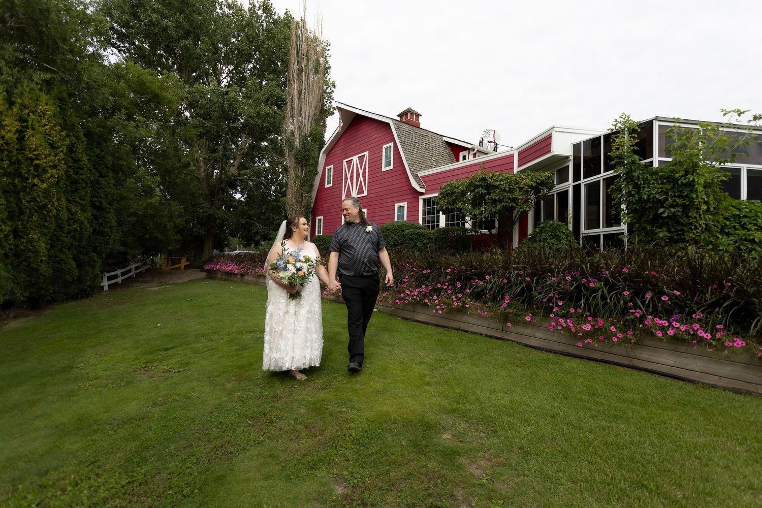 Bride and groom walk hand-in-hand on lush green lawn beside a charming red barn wedding venue.