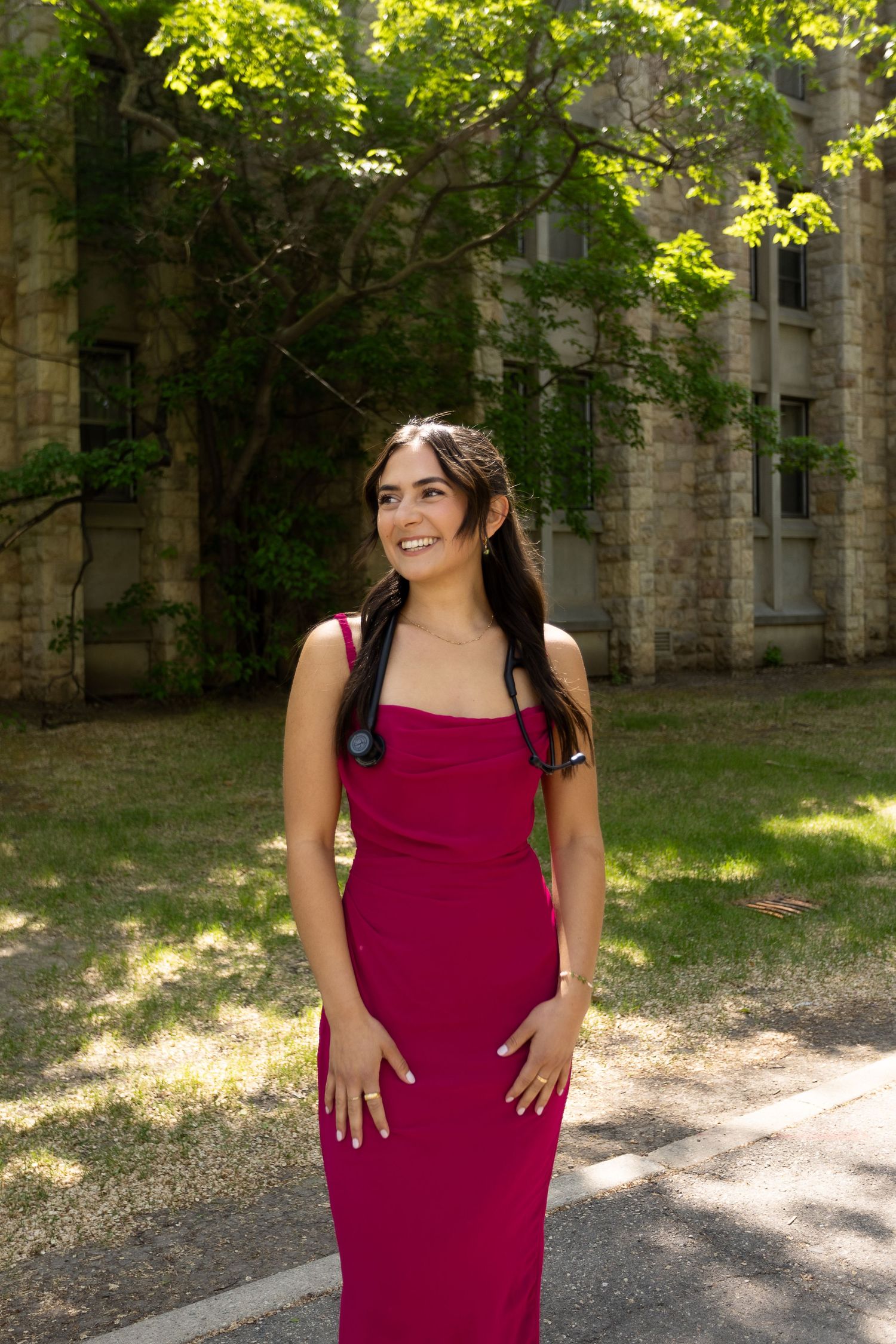 Smiling woman in magenta dress with stethoscope posing outdoors near stone building.
