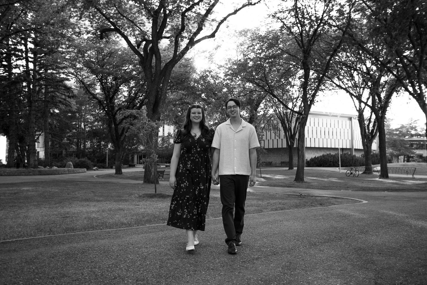 A couple walks hand in hand through a tree-lined park path in a black and white photo.