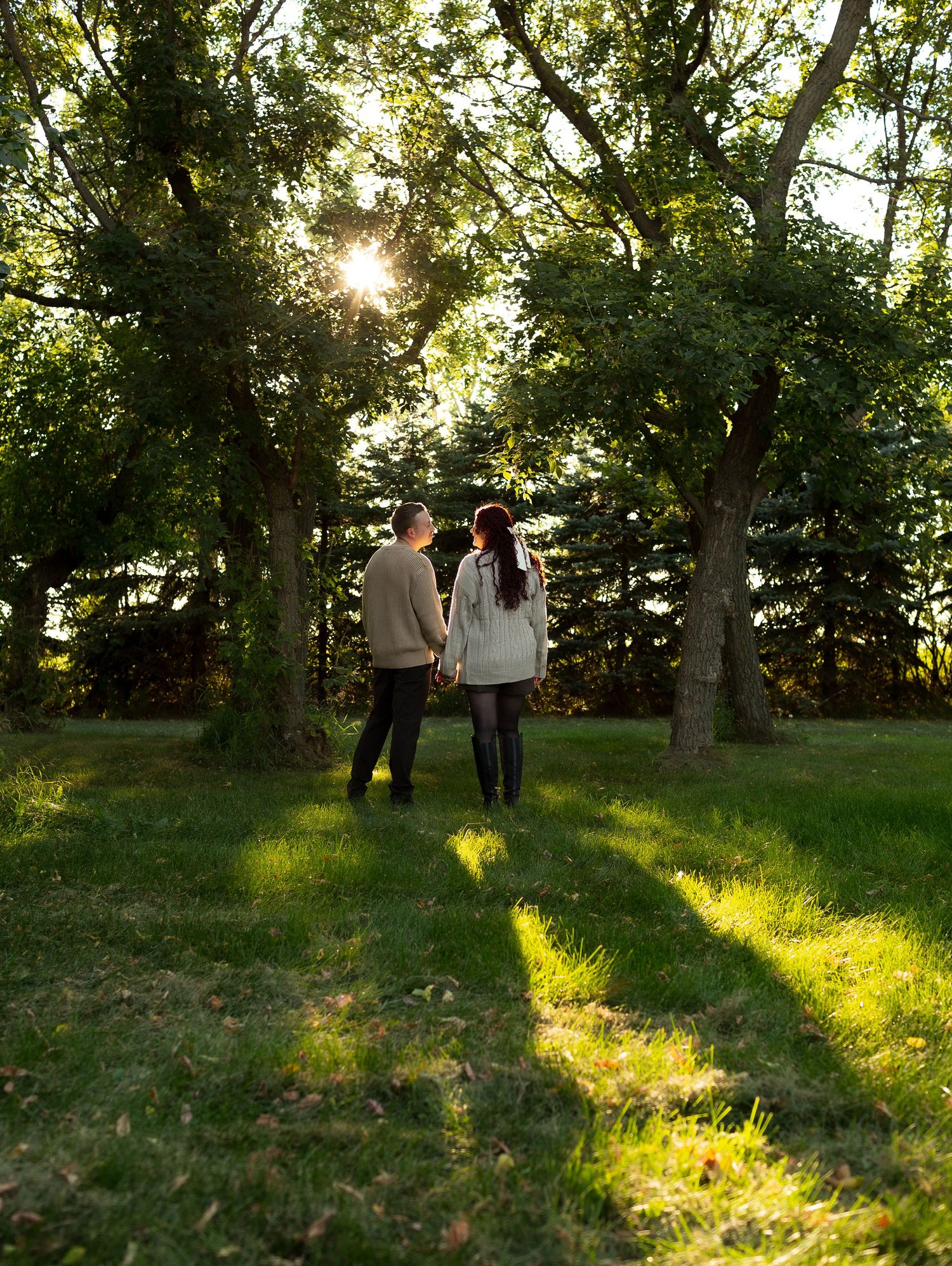 Two people walk through a sunlit park with long shadows cast by trees in late afternoon light.