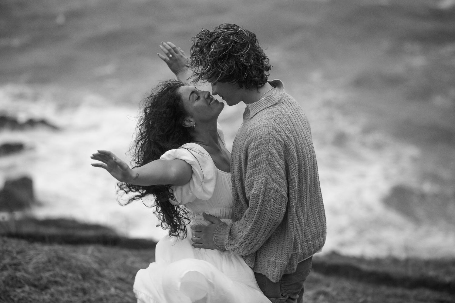 A black and white photo capturing a romantic embrace by the ocean with waves crashing in the background near the coast of BC.