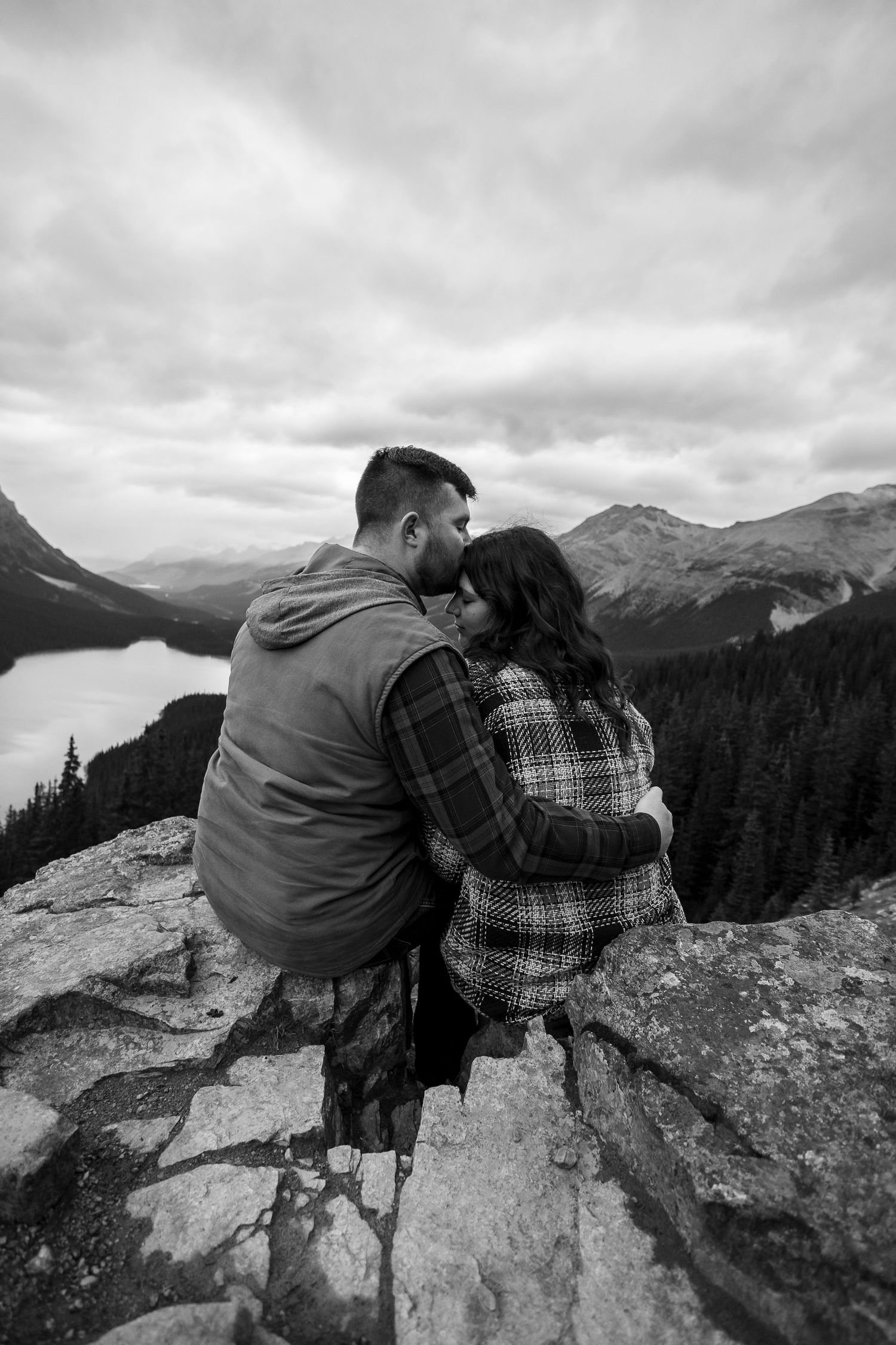 Couple embracing on a rocky mountain overlook with a lake and forest valley below in black and white.