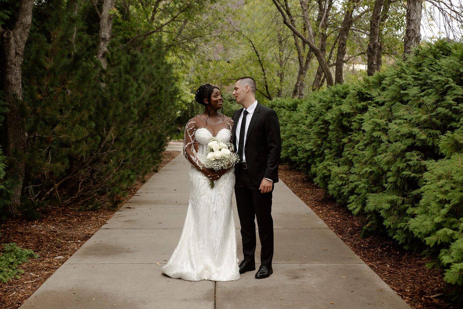 Bride and groom share a romantic moment on a tree-lined path, bride in white gown holding bouquet.