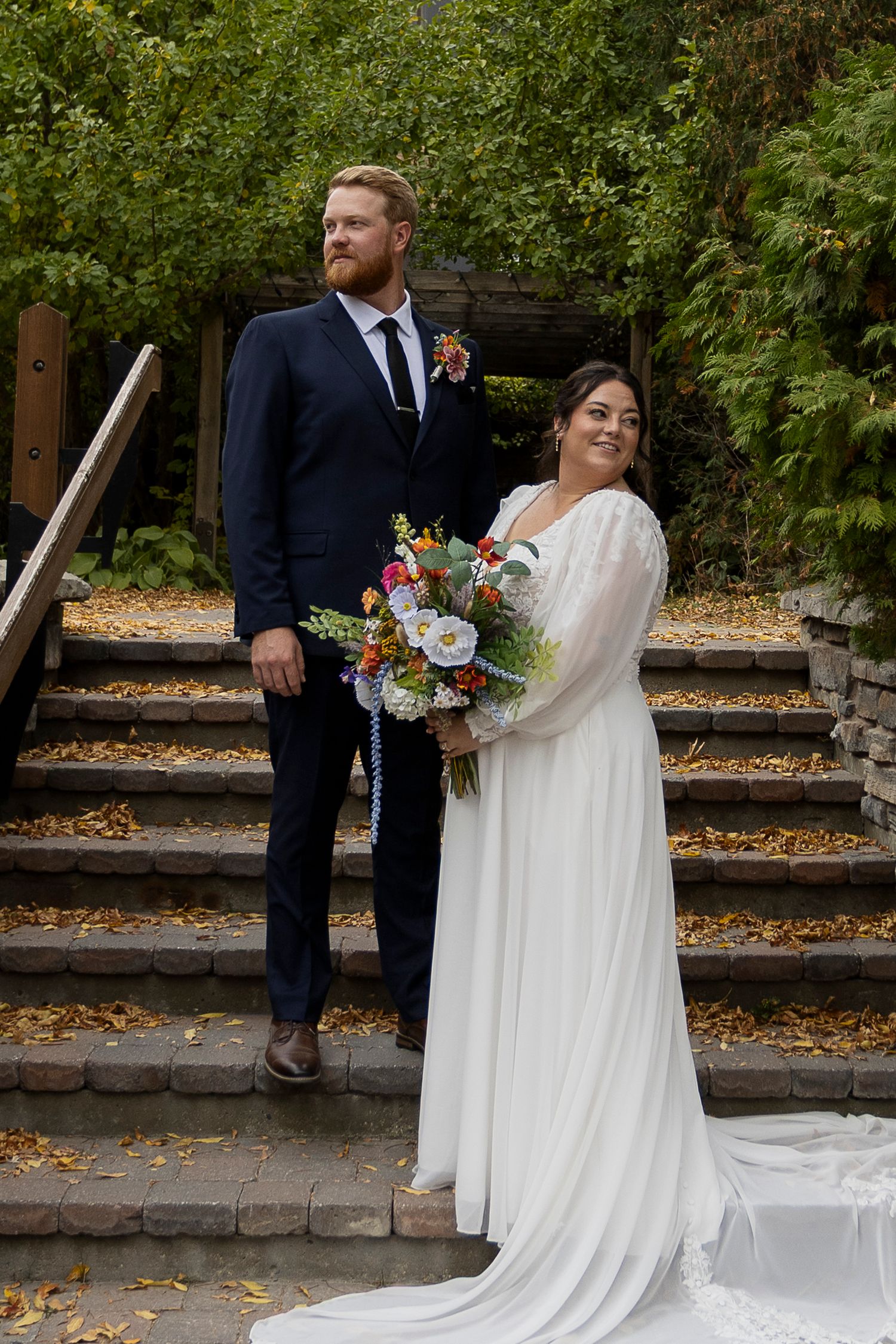 Bride in white gown holding colorful bouquet stands with groom in navy suit on stone steps surrounded by autumn foliage.