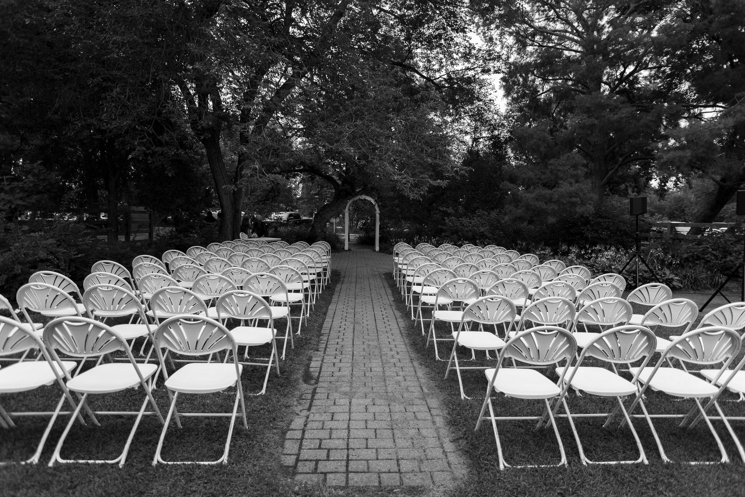 Empty white ceremony chairs lined up on a stone path surrounded by trees at the Forestry Farm in Saskatoon, Saskatchewan.