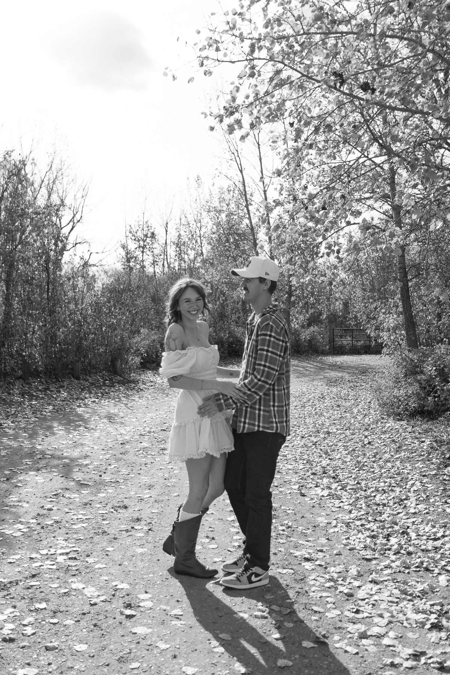 A couple dancing on a leaf-covered path surrounded by autumn trees in black and white.