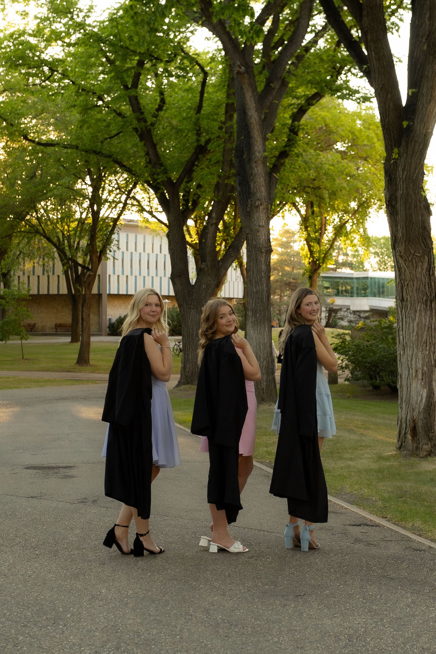 Three women in black graduation gowns pose outdoors on a tree-lined campus path during golden hour.