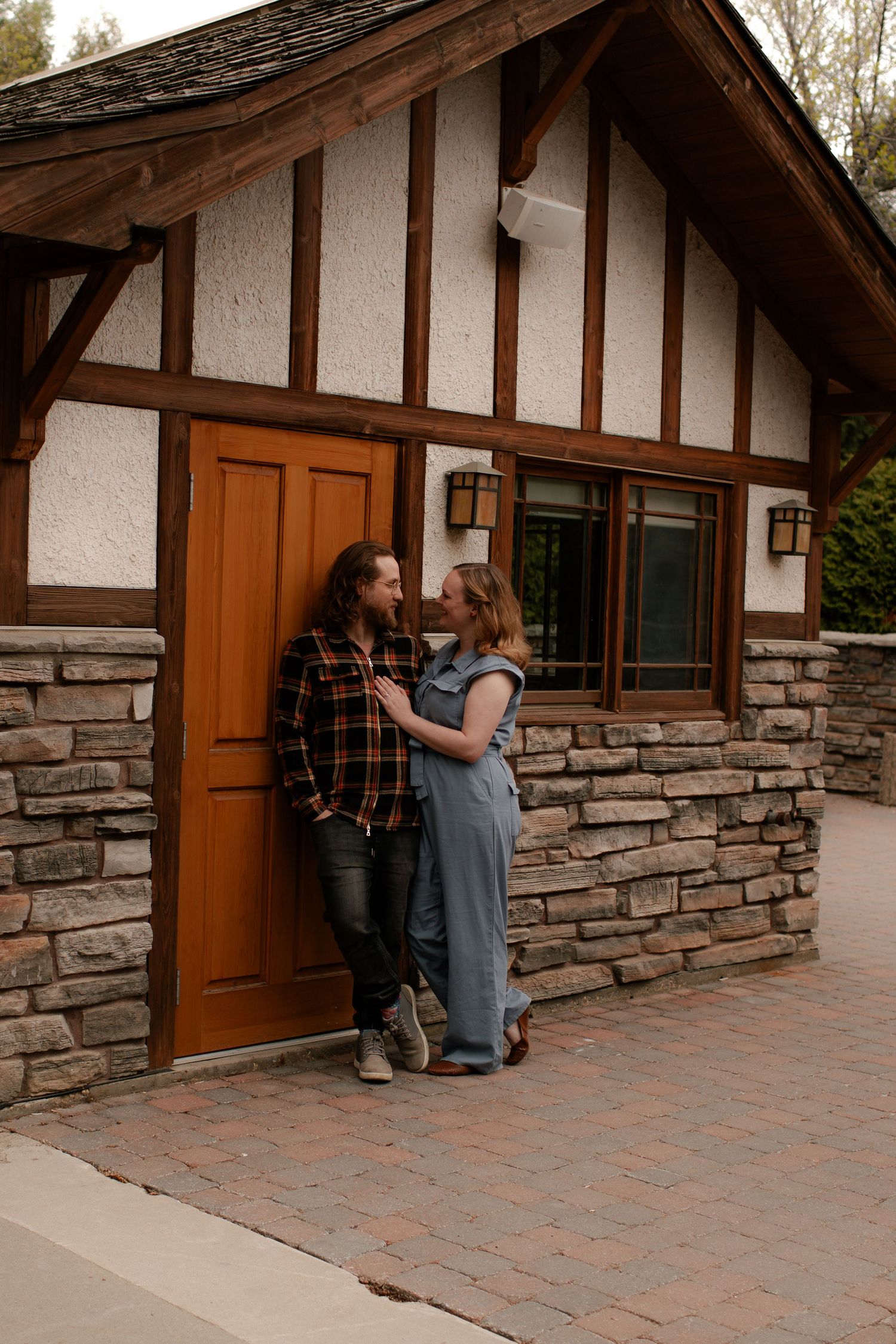 Couple in casual attire sharing a romantic moment outside a Tudor-style stone building with a wooden door.