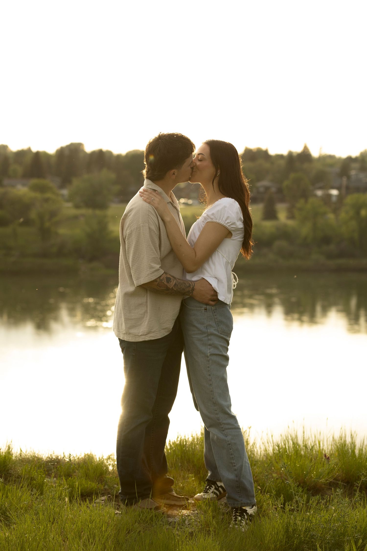 Couple sharing a romantic kiss by a serene lake at golden hour, surrounded by lush green grass and trees.