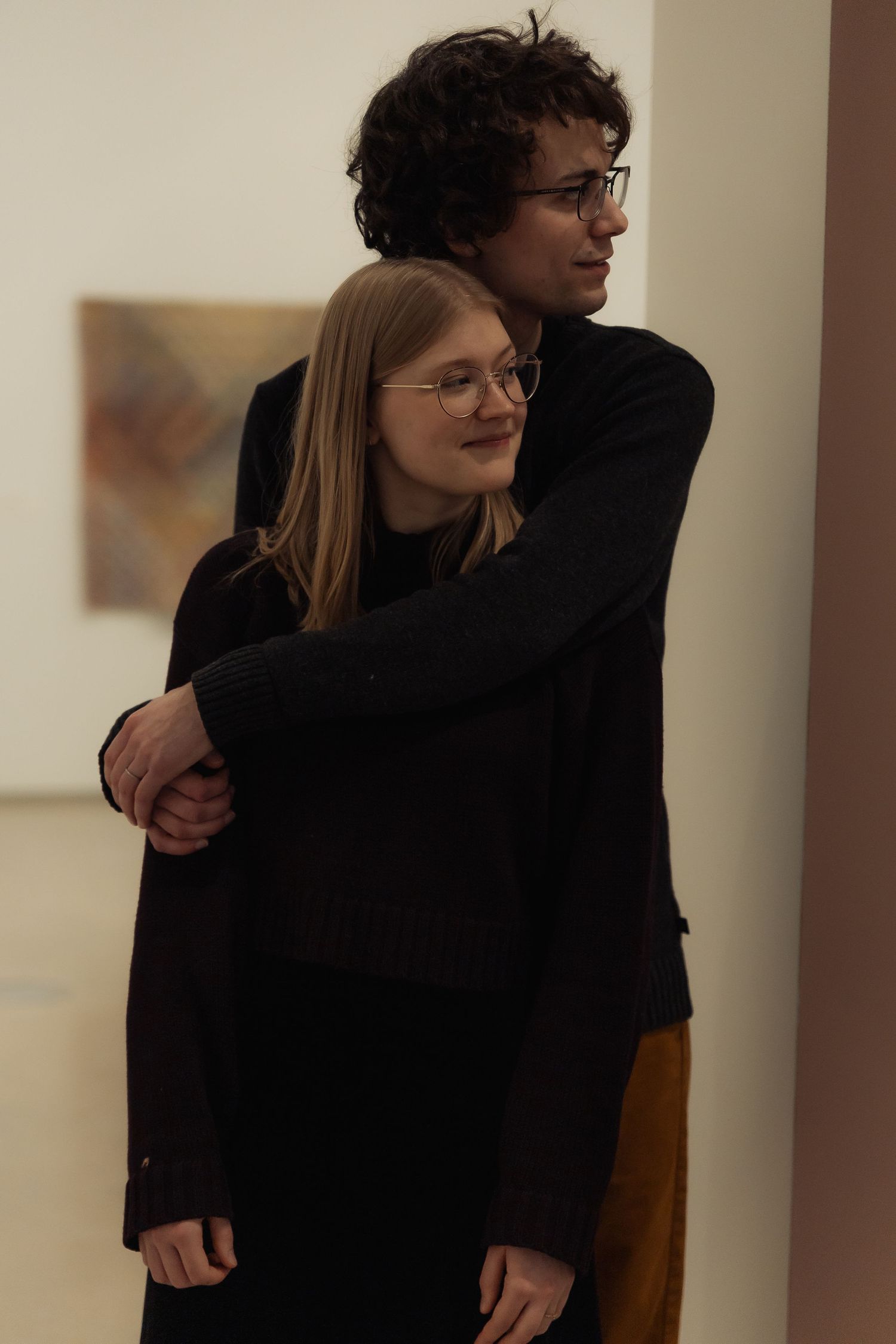 Intimate indoor portrait of a couple embracing while wearing dark clothing at art gallery.