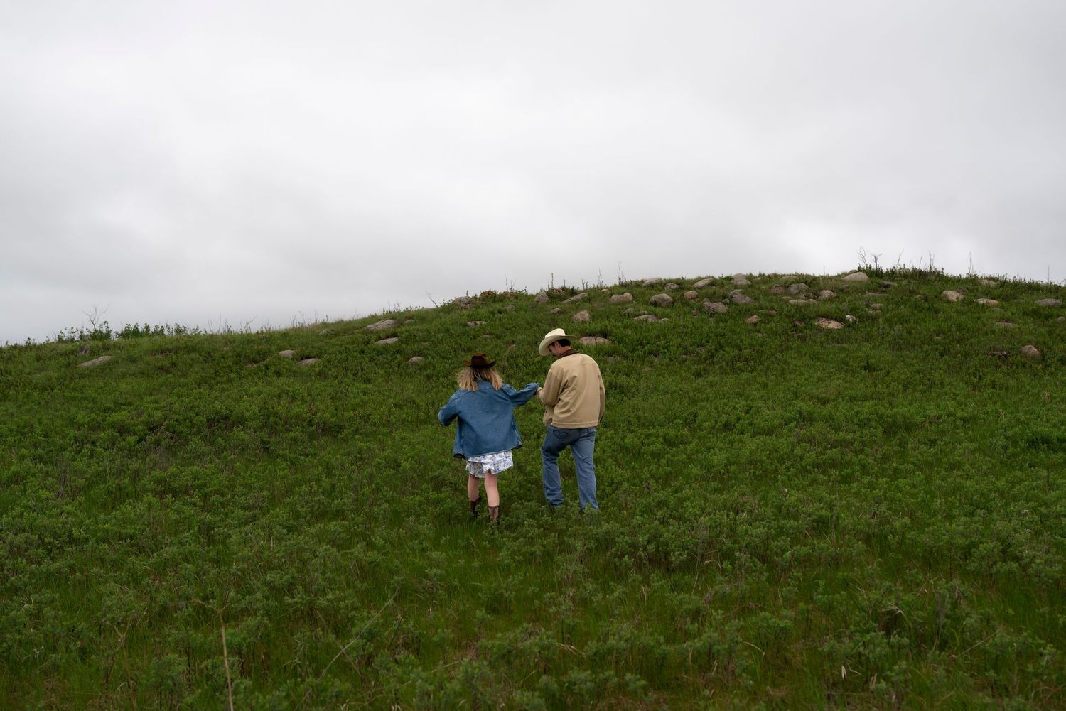 Two people hiking up a lush green hillside on a cloudy day, one guiding the other up the grassy slope.