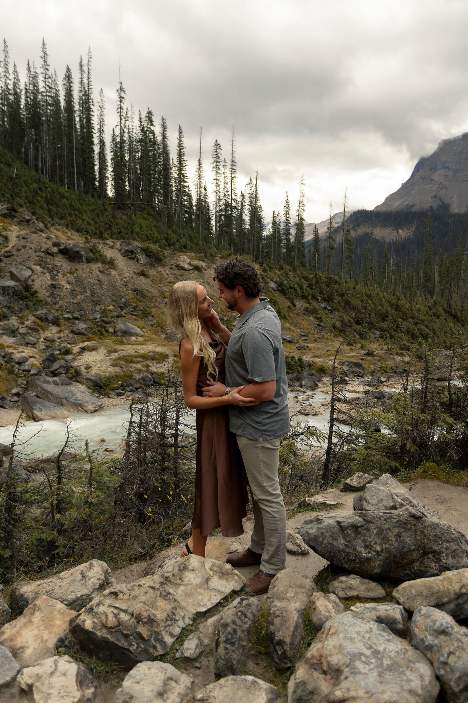 A couple shares a romantic moment on rocky mountain terrain with evergreen trees in the background.