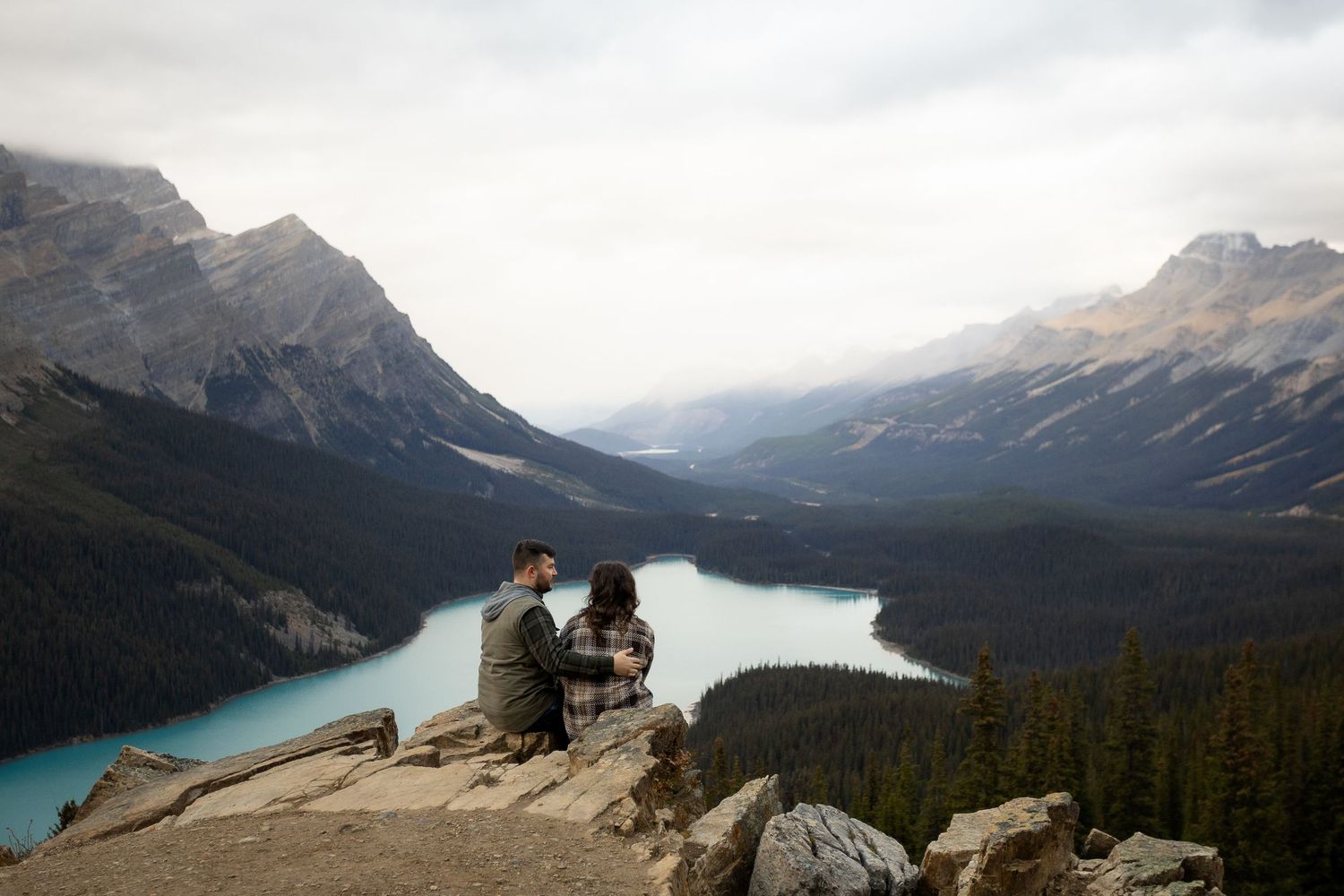 Couple sitting on rocky cliff overlooking turquoise Peyto Lake surrounded by mountains and pine forests in Banff, Canada.