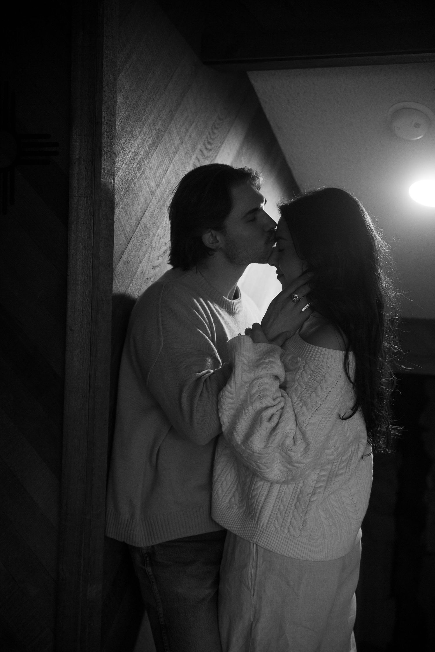 Black and white photo of a romantic couple sharing a tender kiss indoors, bathed in soft backlight.