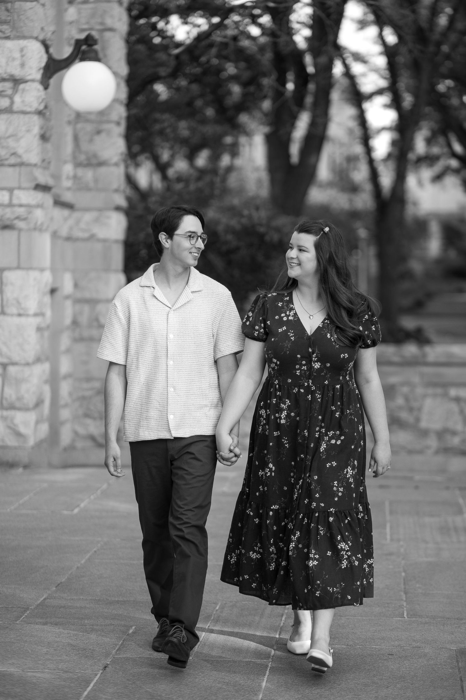 A couple walks hand-in-hand outdoors, smiling at each other, in a black and white photo near stone architecture.