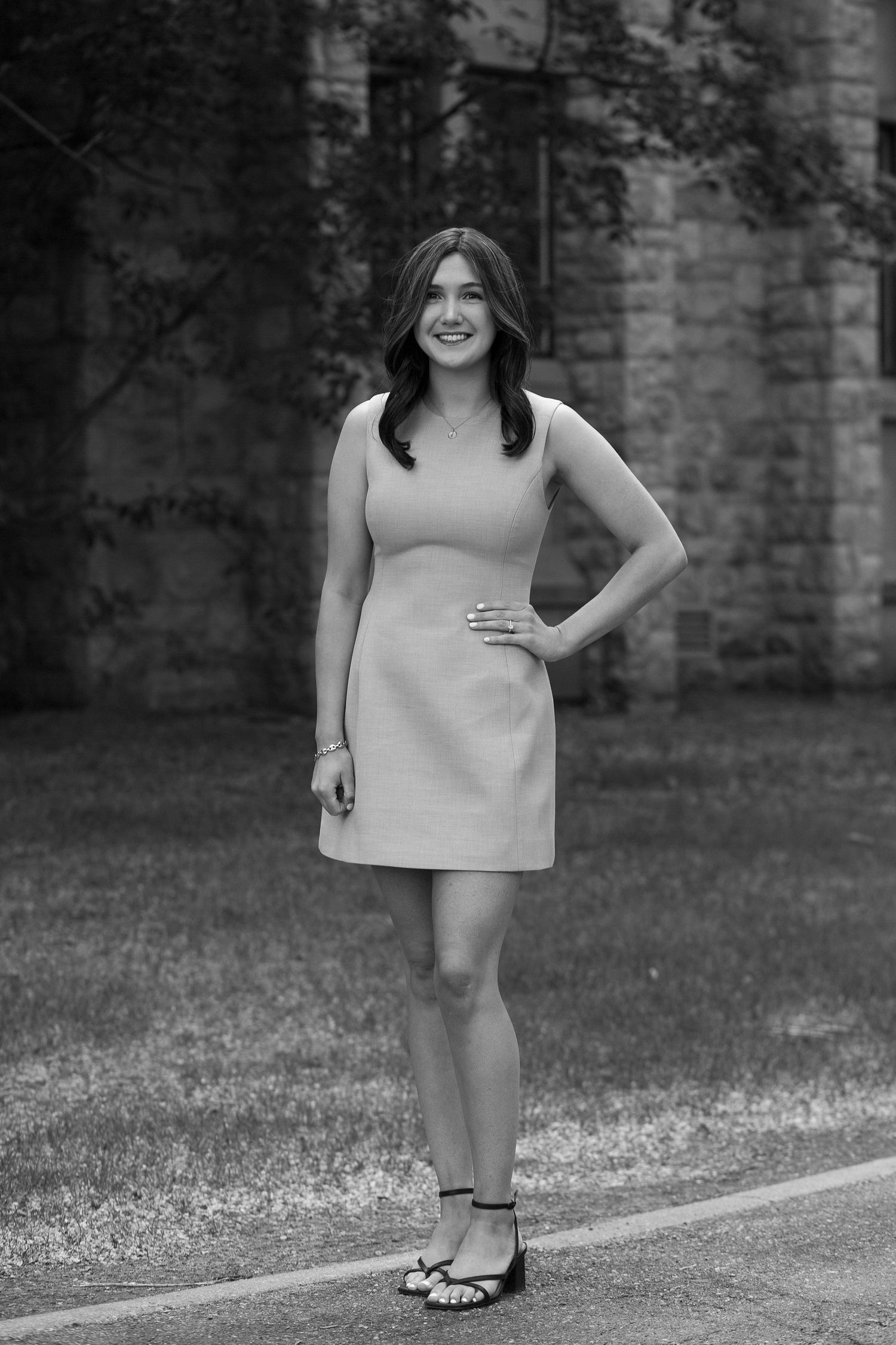Black and white photo of a smiling young woman in a short dress posing outdoors near a stone wall.