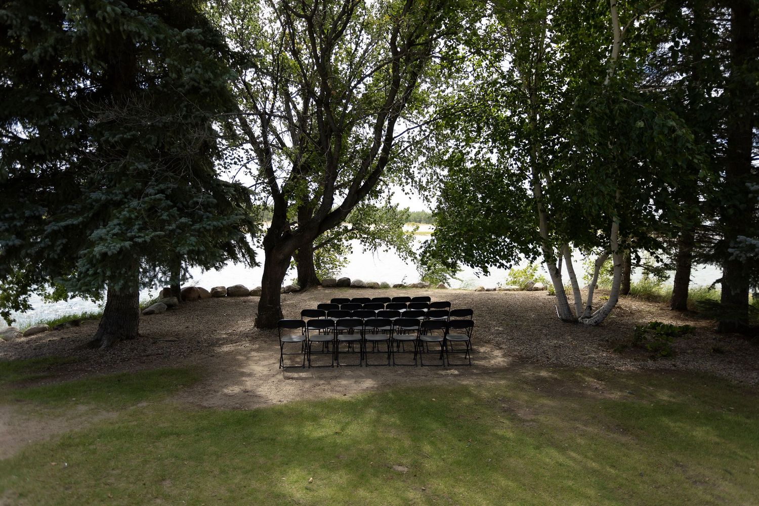 Rows of black chairs arranged outdoors under large trees for an elegant garden ceremony near a serene waterfront.