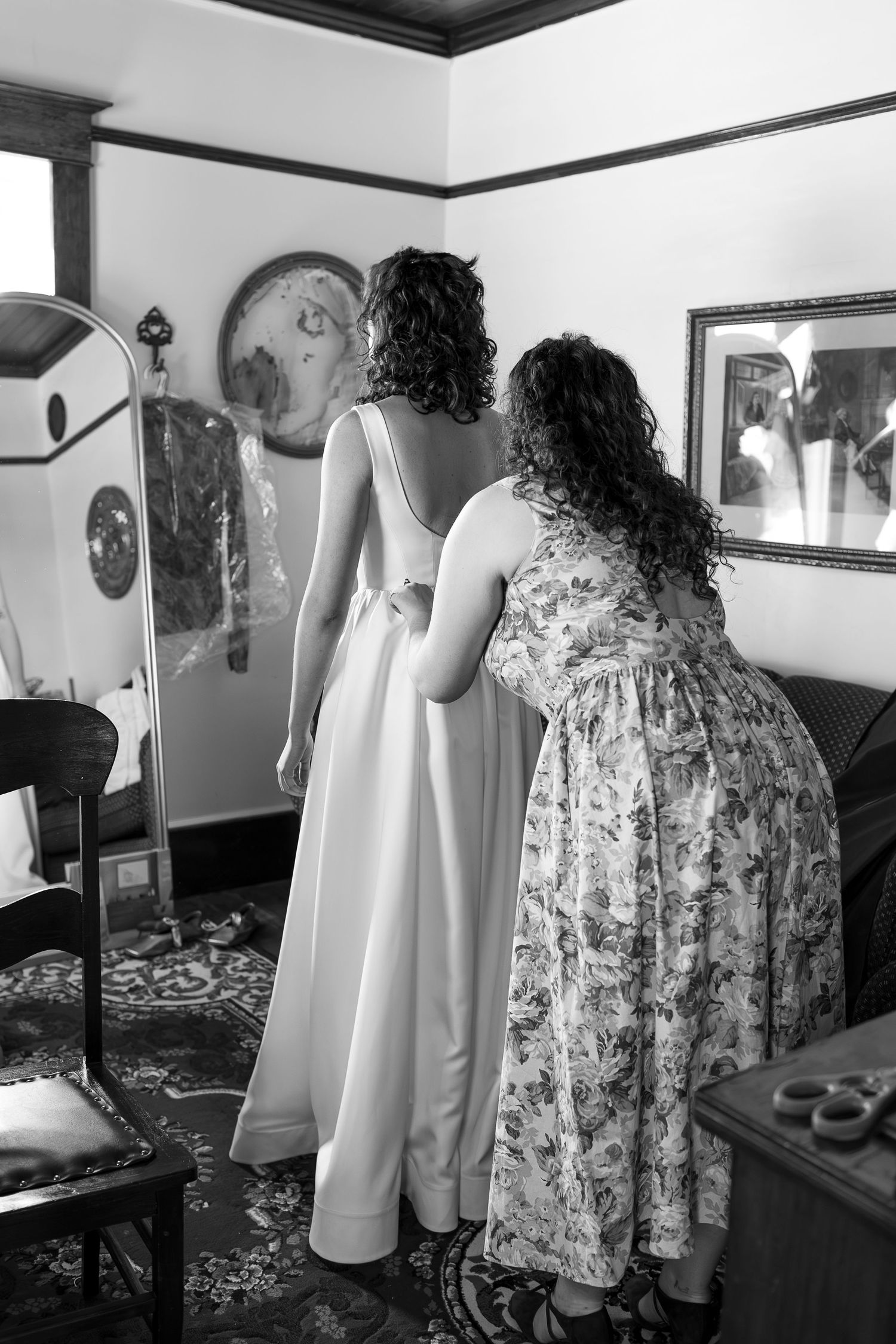 Black and white photo of a bride in a white gown being helped by a woman in a floral dress in an elegant room.