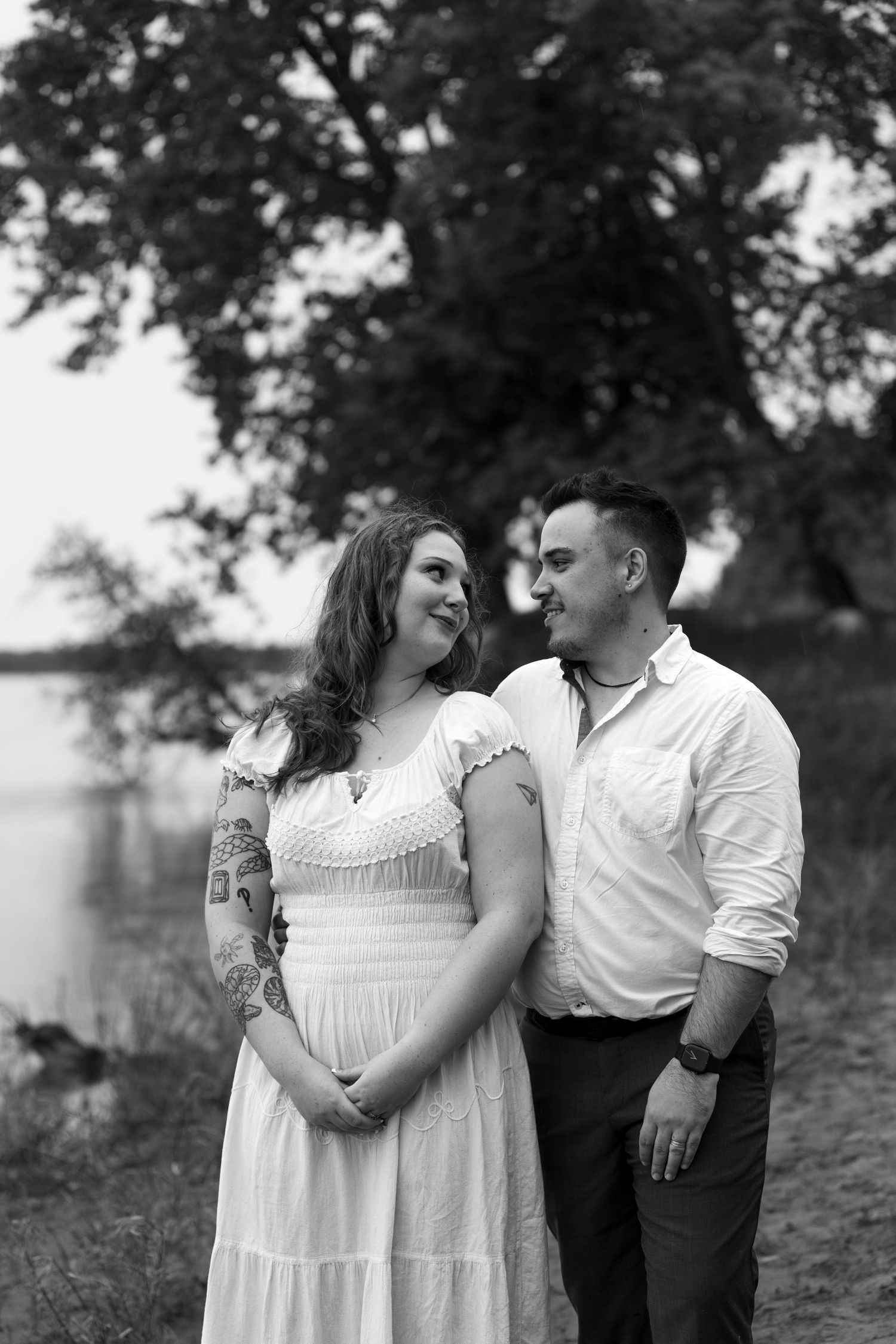 Black and white photo of a couple in white dress and shirt standing by a lake among trees.