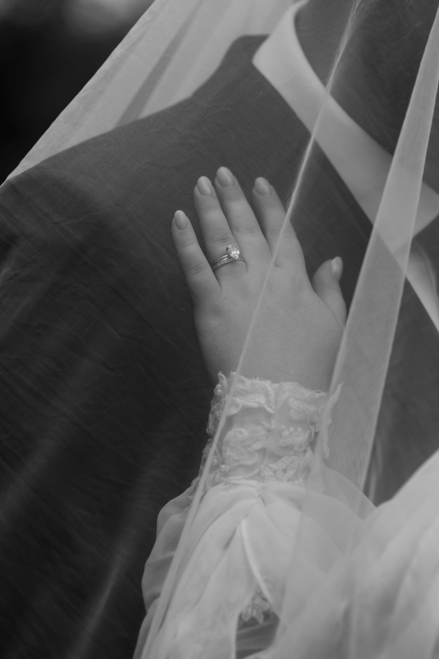 Black and white photo of a bride's hand wearing a diamond ring, draped in elegant lace sleeve and sheer wedding veil.