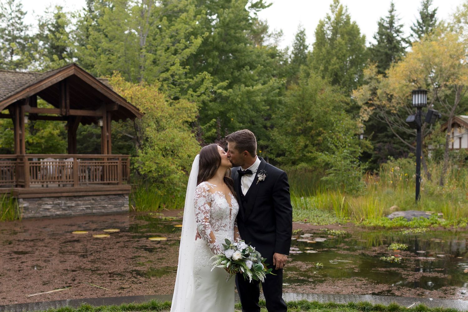 Bride and groom share a kiss outdoors near a rustic gazebo and pond surrounded by lush green trees.