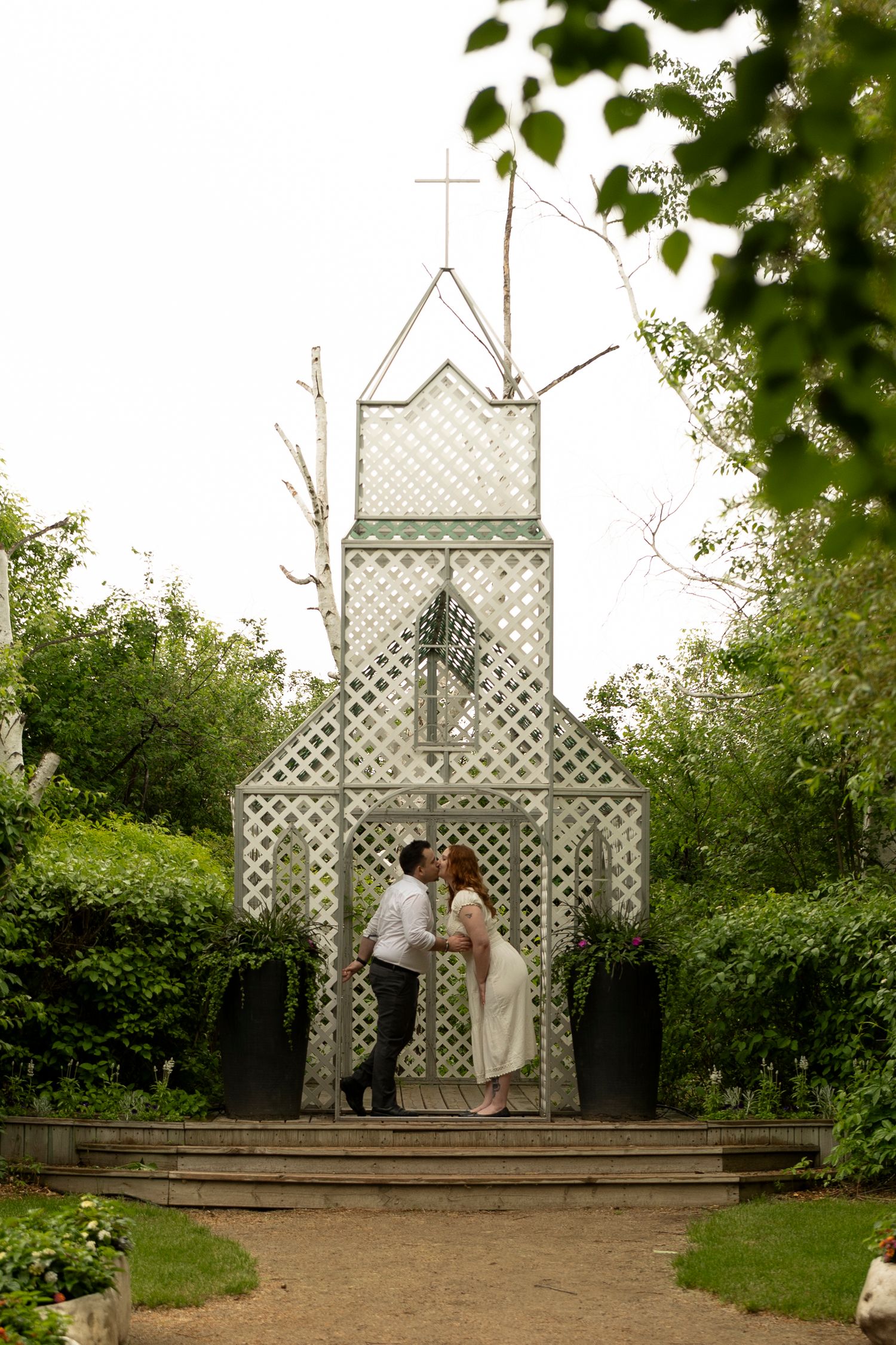 Couple sharing a kiss in front of a charming white lattice chapel surrounded by lush greenery.