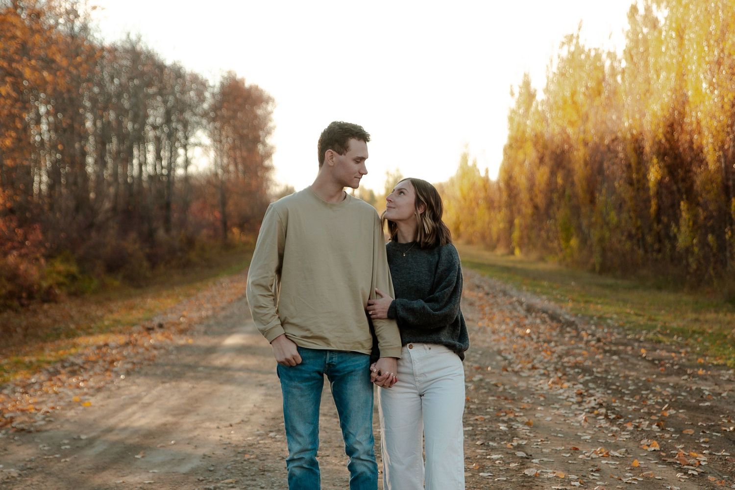 Couple holding hands on a scenic autumn dirt road surrounded by colorful fall foliage at golden hour.