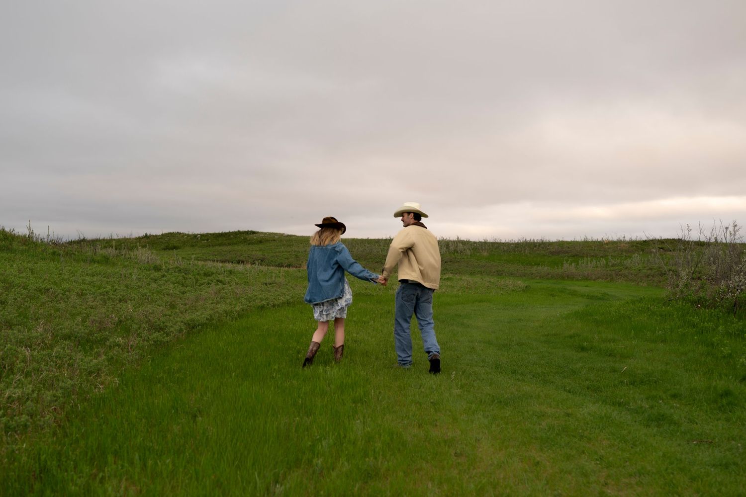 Two people in cowboy hats hold hands on a lush green field under an overcast sky.