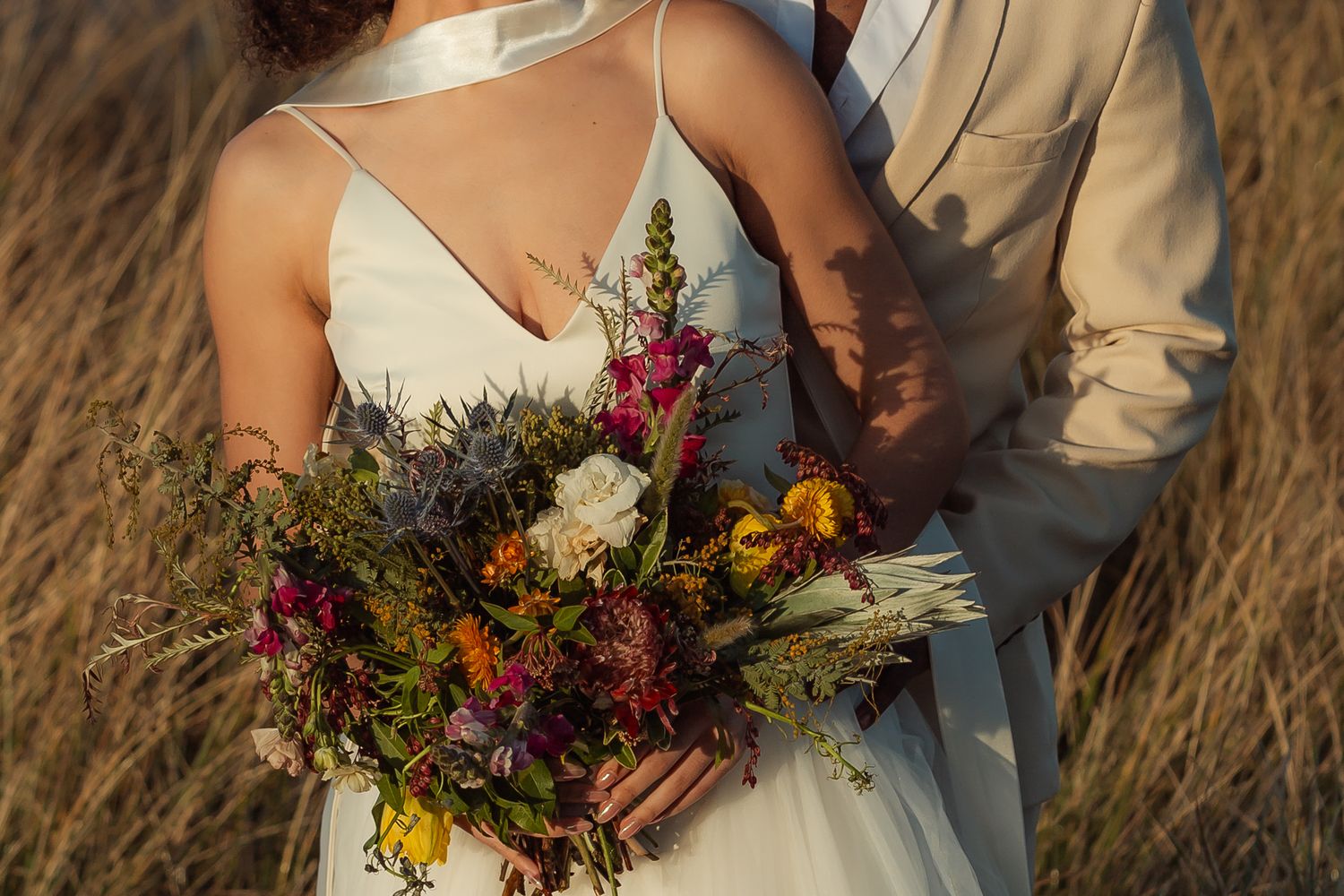 Close-up of dried flower wedding bouquet with colorful wildflowers held against white dress.